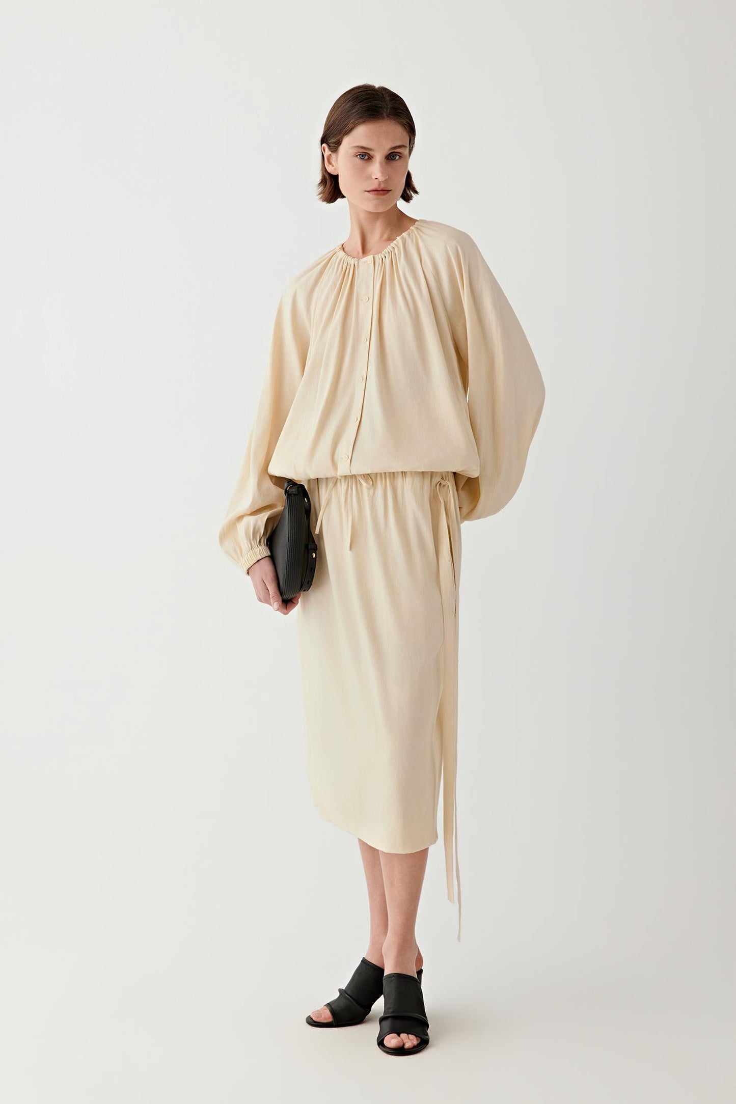Full-length front view of a woman standing facing forward wearing a sand-colored linen blend midi skirt, paired with a matching top, highlighting the fluid A-line silhouette, lightweight structure and soft drape of the fabric falling below the knee
