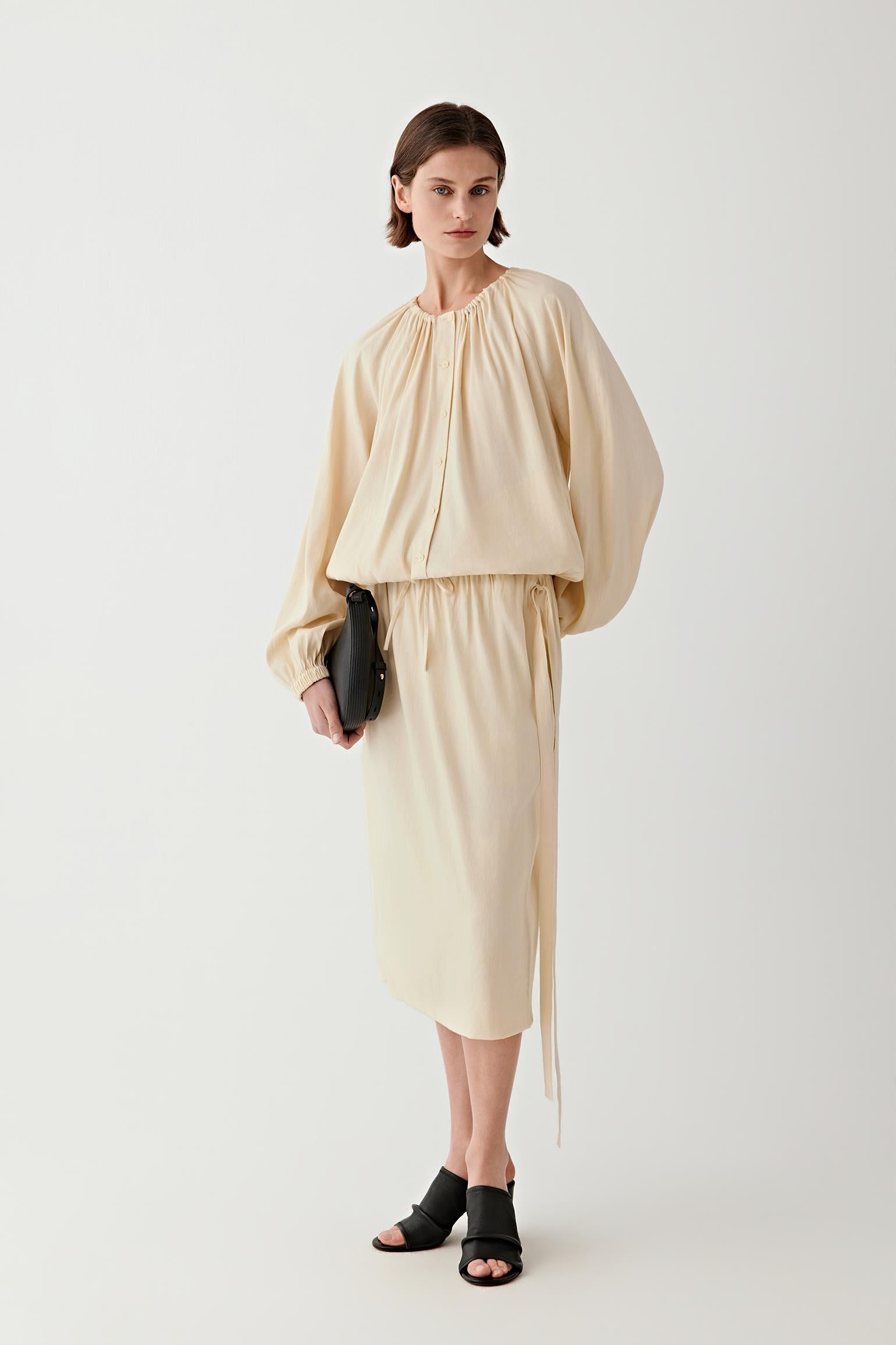 Full-length front view of a woman standing facing forward wearing a sand-colored linen blend midi skirt, paired with a matching top, highlighting the fluid A-line silhouette, lightweight structure and soft drape of the fabric falling below the knee