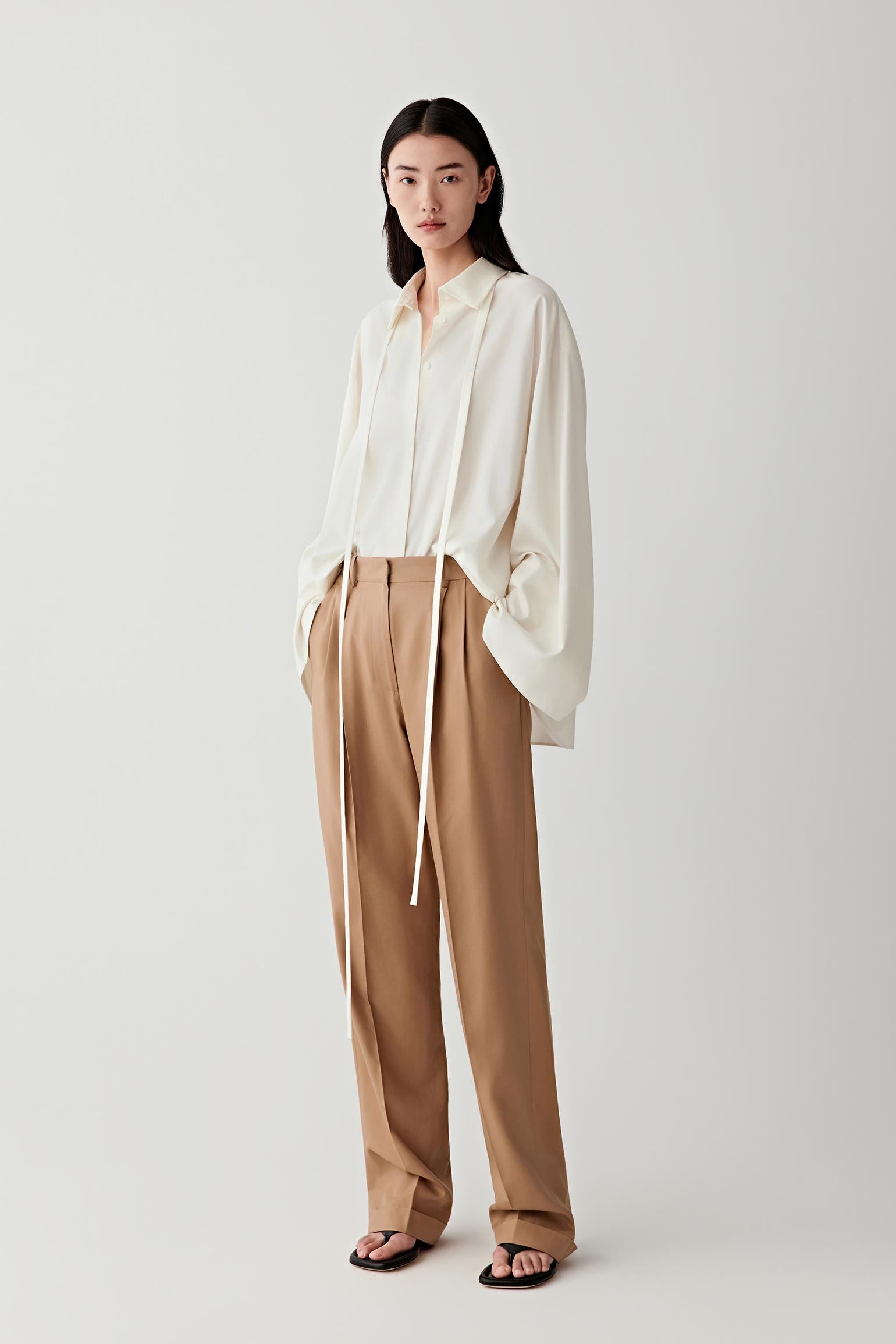 Full-length front view of a woman wearing camel straight-leg trousers in spun silk poplin, highlighting the fluid drape, smooth surface and soft tonal variation