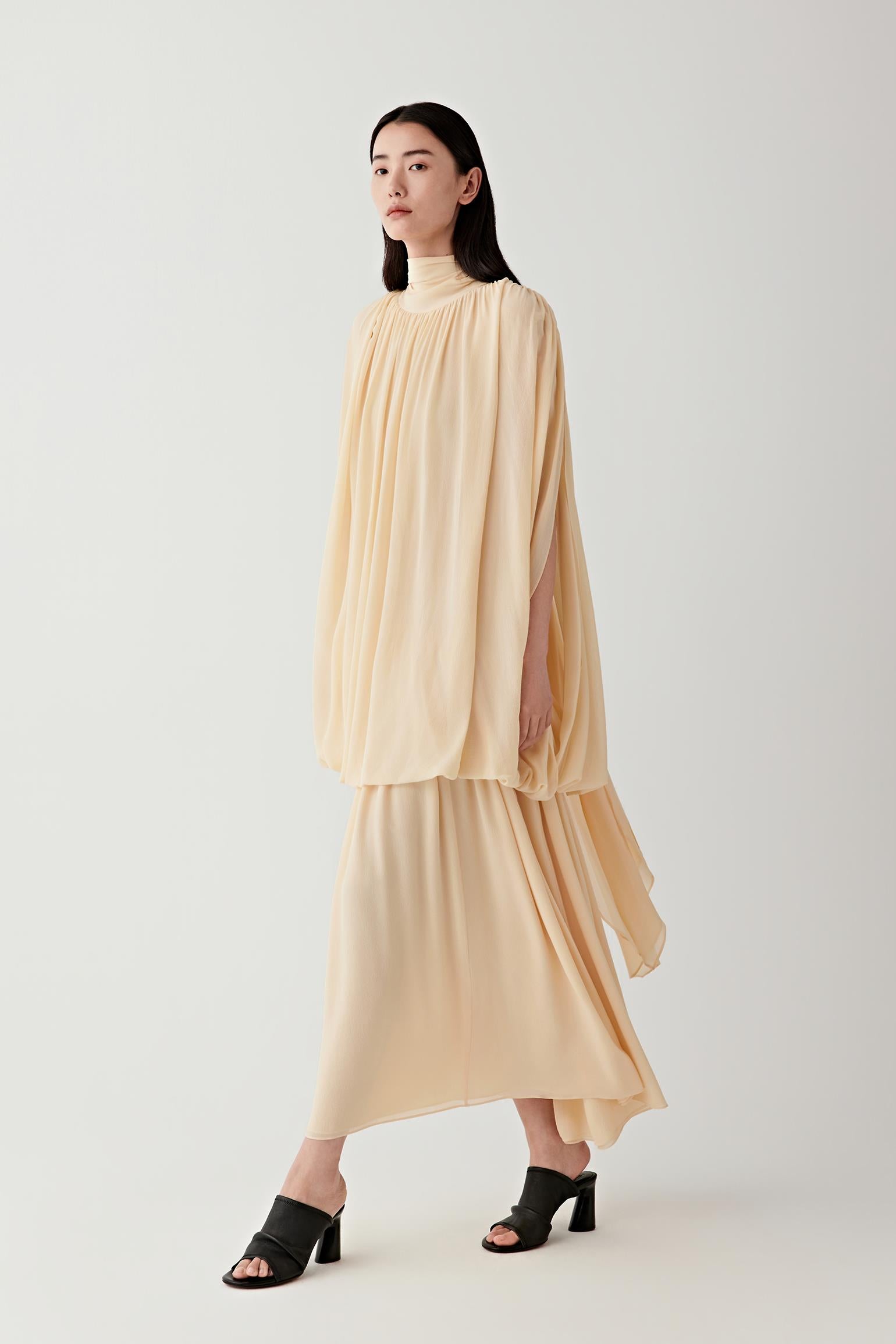 Full-length front view of a woman walking slightly forward wearing a pale lemon yellow silk georgette long dress, highlighting the elongated silhouette, fluid drape and softly matte, lightweight texture, with layered panels creating gentle movement along the length