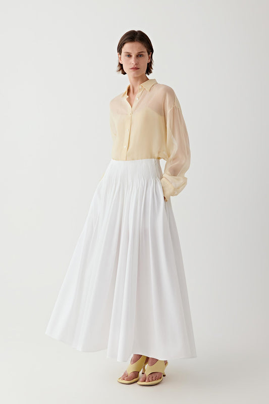 Full-length front view showing the maxi-length silhouette shaped by precise pleats and a high waist construction. The structured cotton poplin maintains volume and definition through the length of the skirt.