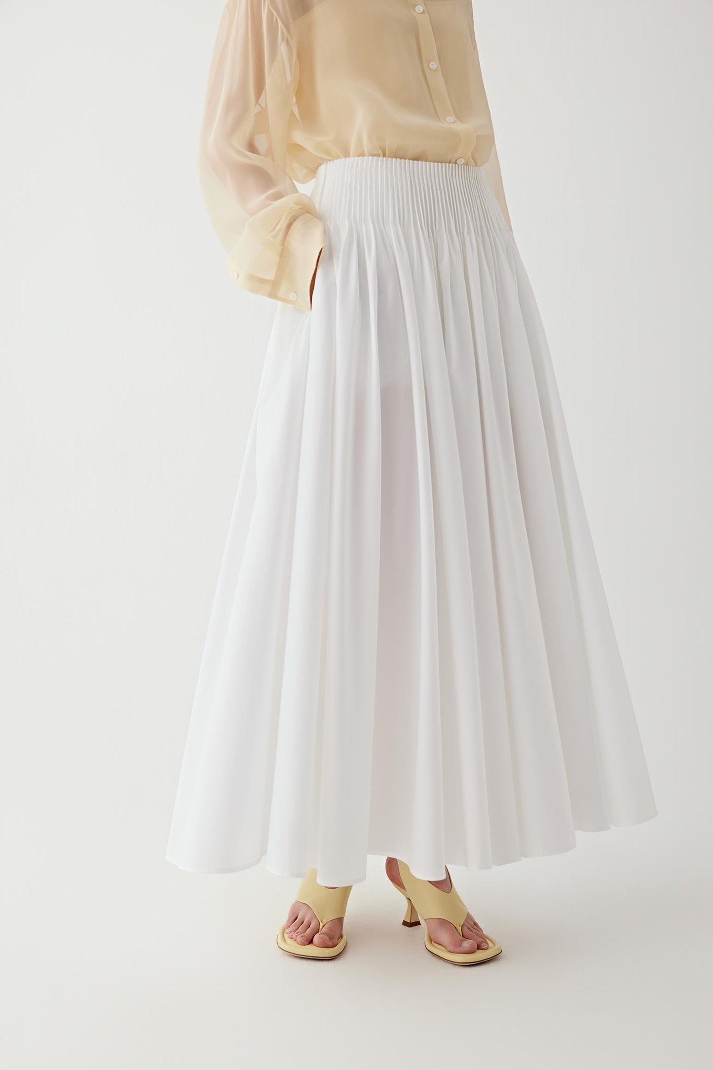 Front view of a woman wearing a white pleated maxi skirt in cotton poplin, highlighting the crisp surface, structured drape and matte finish of the fabric. The poplin construction creates a clean and architectural silhouette.