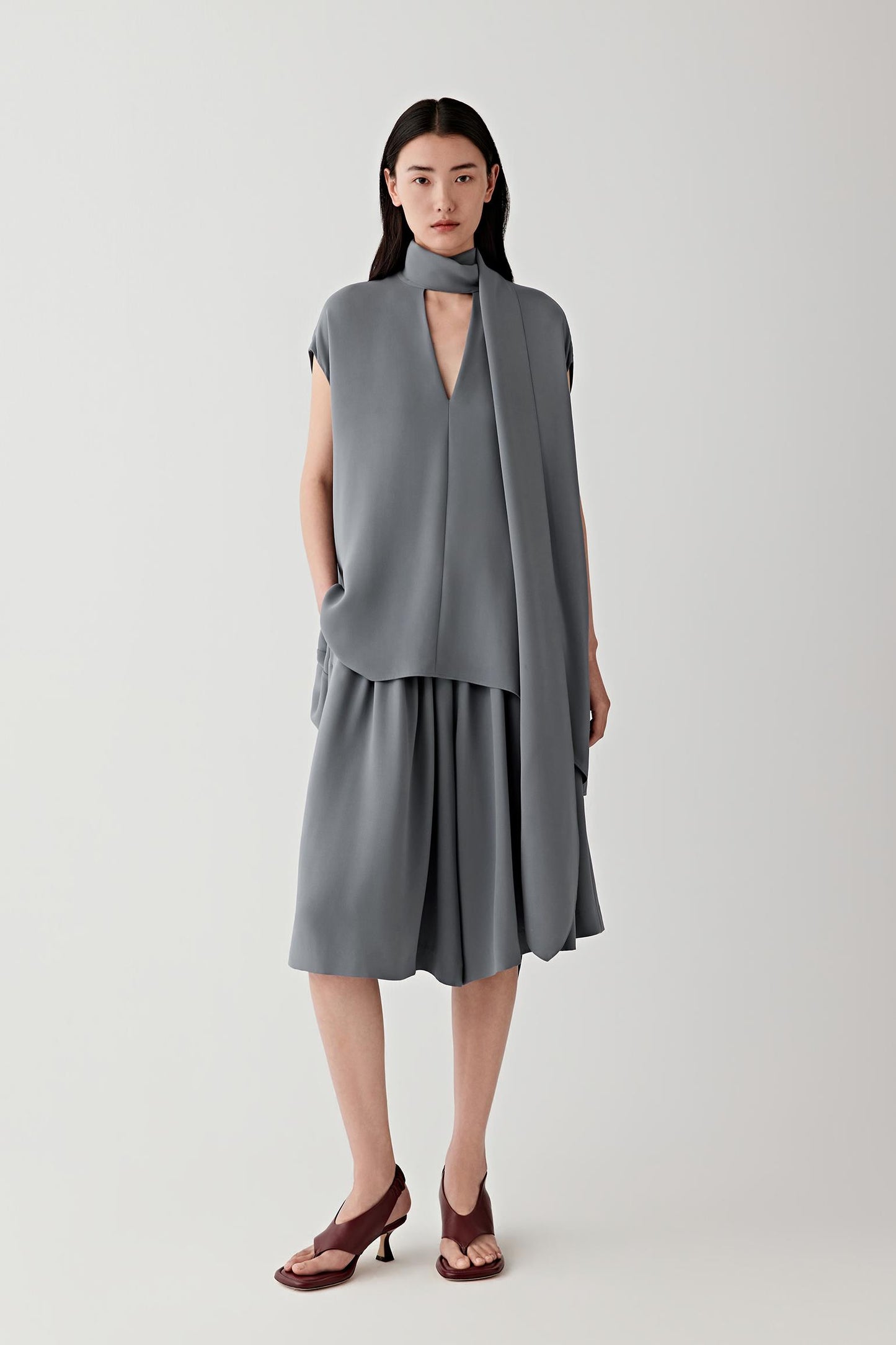 Full-length front view showing the elongated silhouette and wrap-style neckline extending into a front panel construction. The sleeveless cut and fluid silk crepe create soft movement while maintaining clean vertical lines.