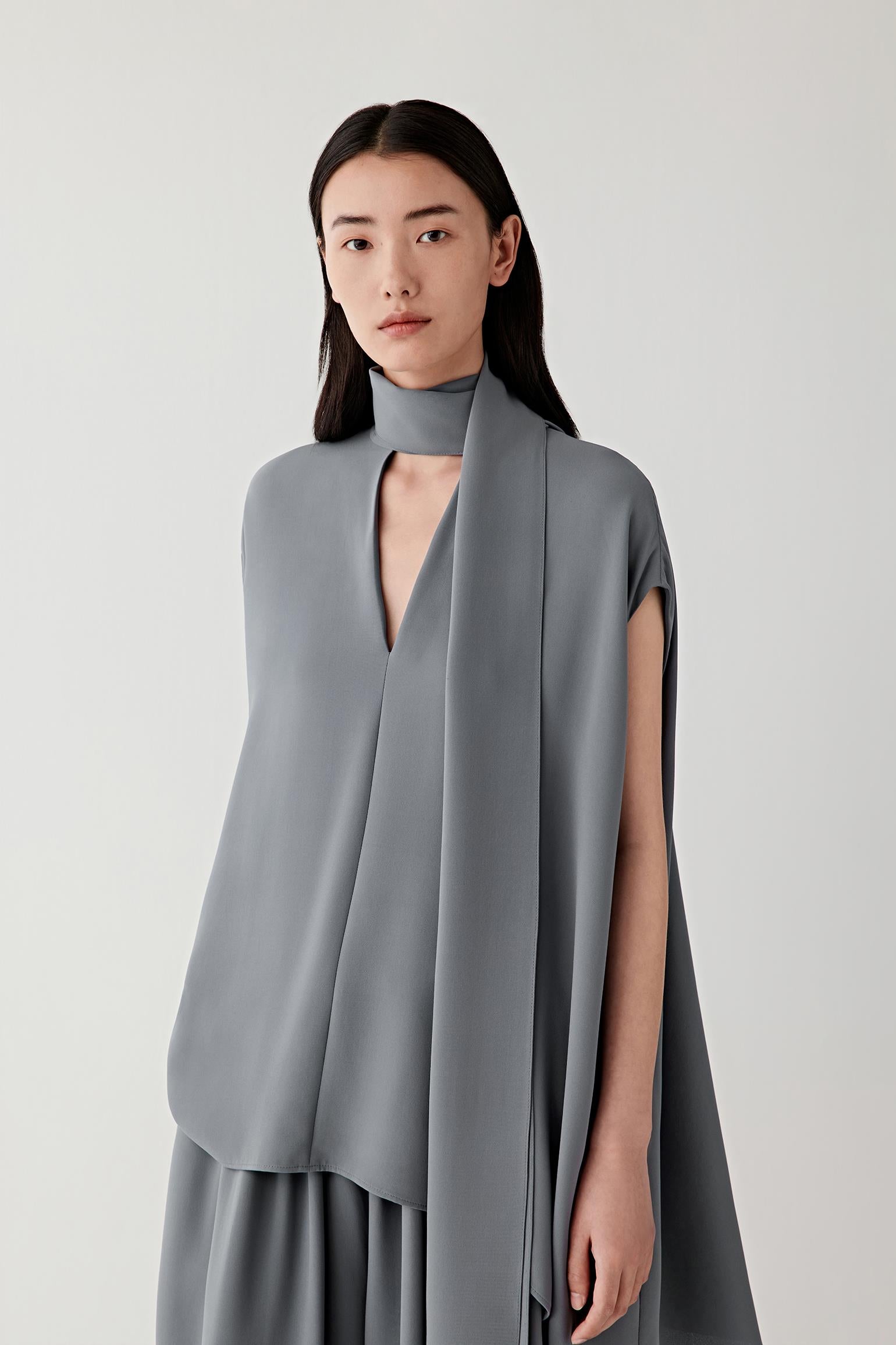 Front view of a woman wearing a grey sleeveless blouse in heavy silk crepe, highlighting the smooth matte surface, soft hand and fluid drape of the fabric. The material reveals subtle tonal depth and a softly structured fall through the silhouette.