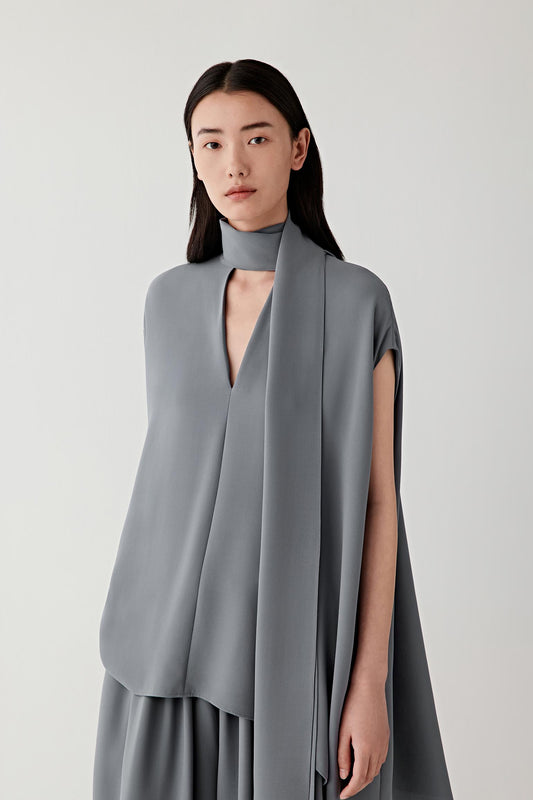 Front view of a woman wearing a grey sleeveless blouse in heavy silk crepe, highlighting the smooth matte surface, soft hand and fluid drape of the fabric. The material reveals subtle tonal depth and a softly structured fall through the silhouette.