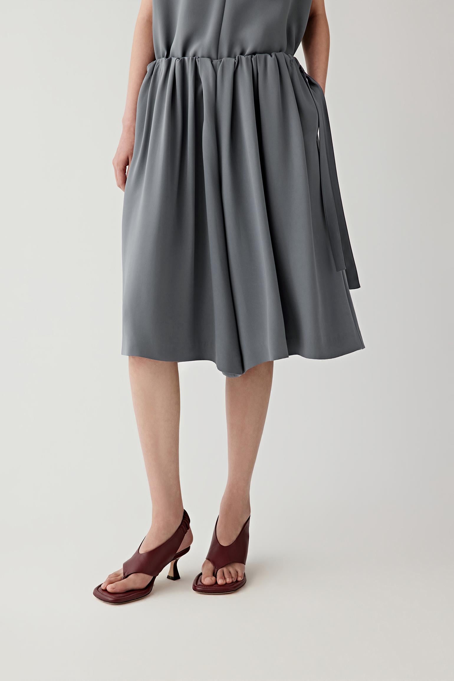 Full-length front view showing the relaxed straight silhouette and elasticated waistband construction. Soft regular pleats emerge from the waistband, creating movement and volume through the garment.