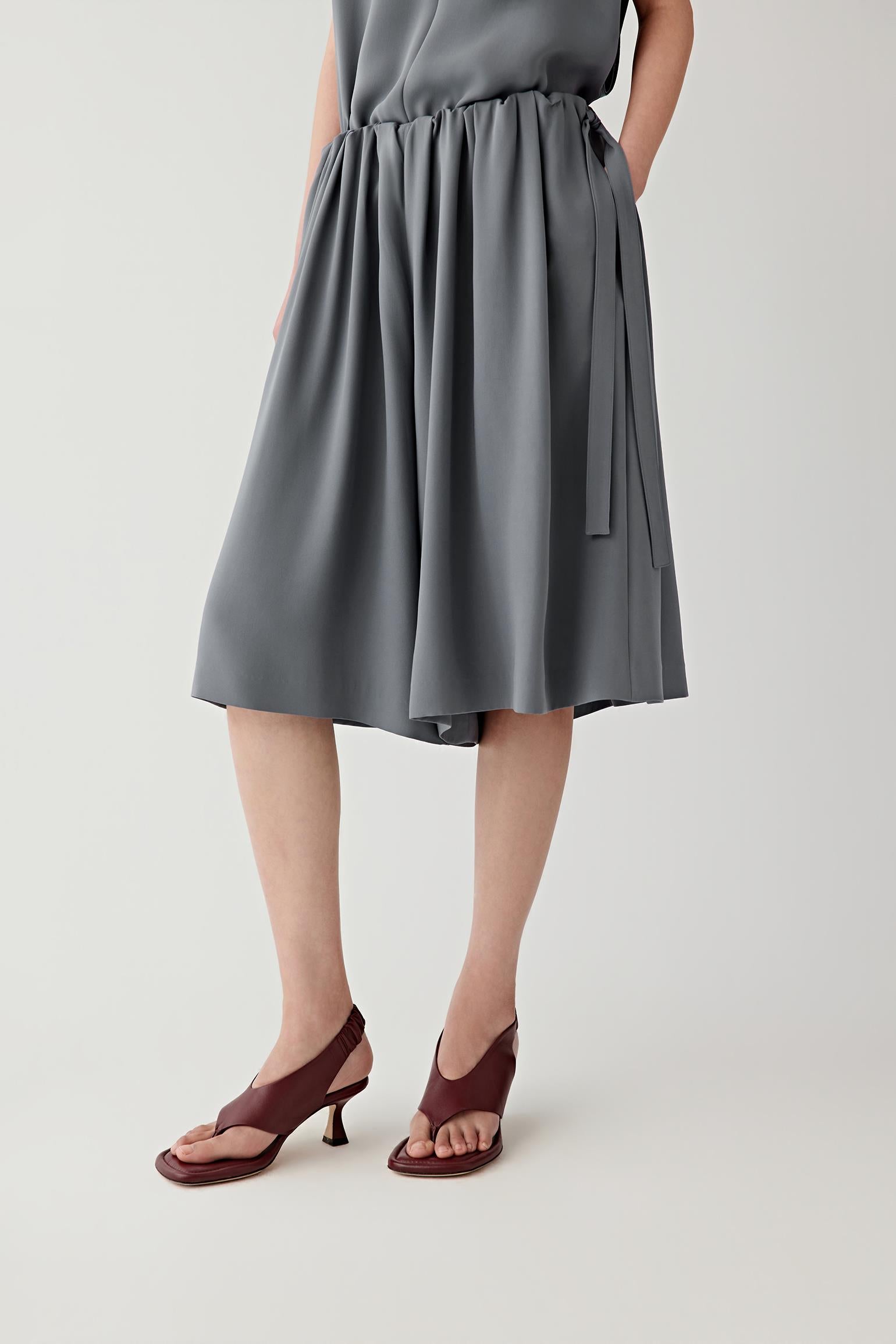 Mid-length front view showing the relaxed straight silhouette and elasticated waistband construction. Soft regular pleats emerge from the waistband, creating movement and volume through the garment.