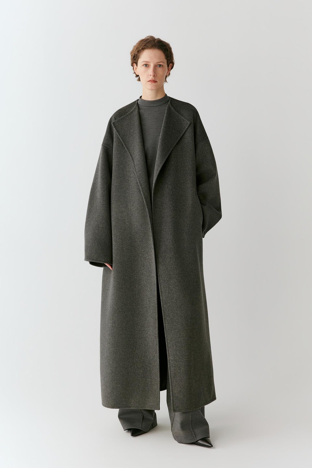A model stands against a light gray studio background wearing a long, oversized dark gray wool coat over a matching dark gray top and wide-leg pants.