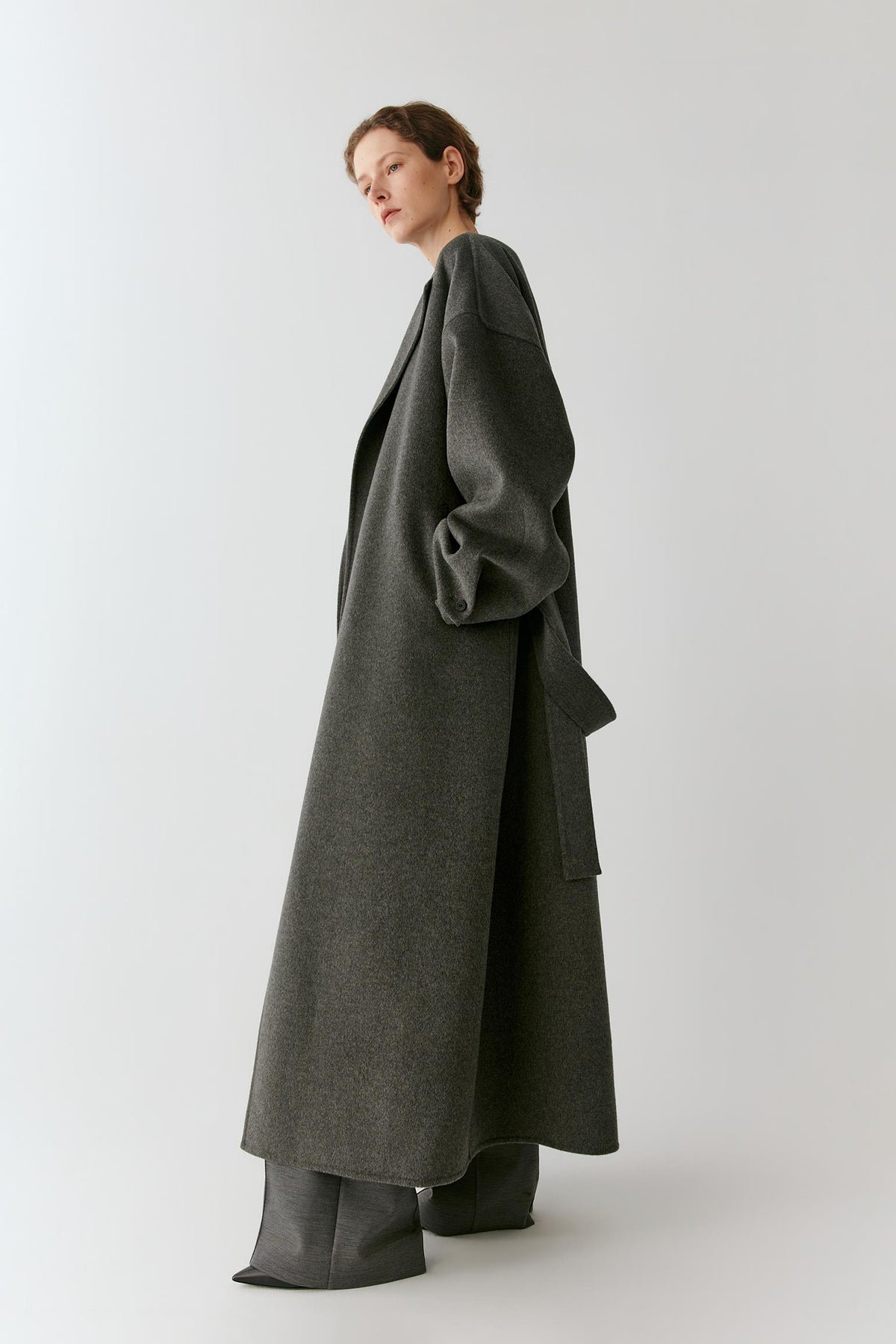 A model wearing a long, dark gray wool-blend coat with oversized draped sleeves and wide-leg trousers, photographed against a neutral light gray studio background.