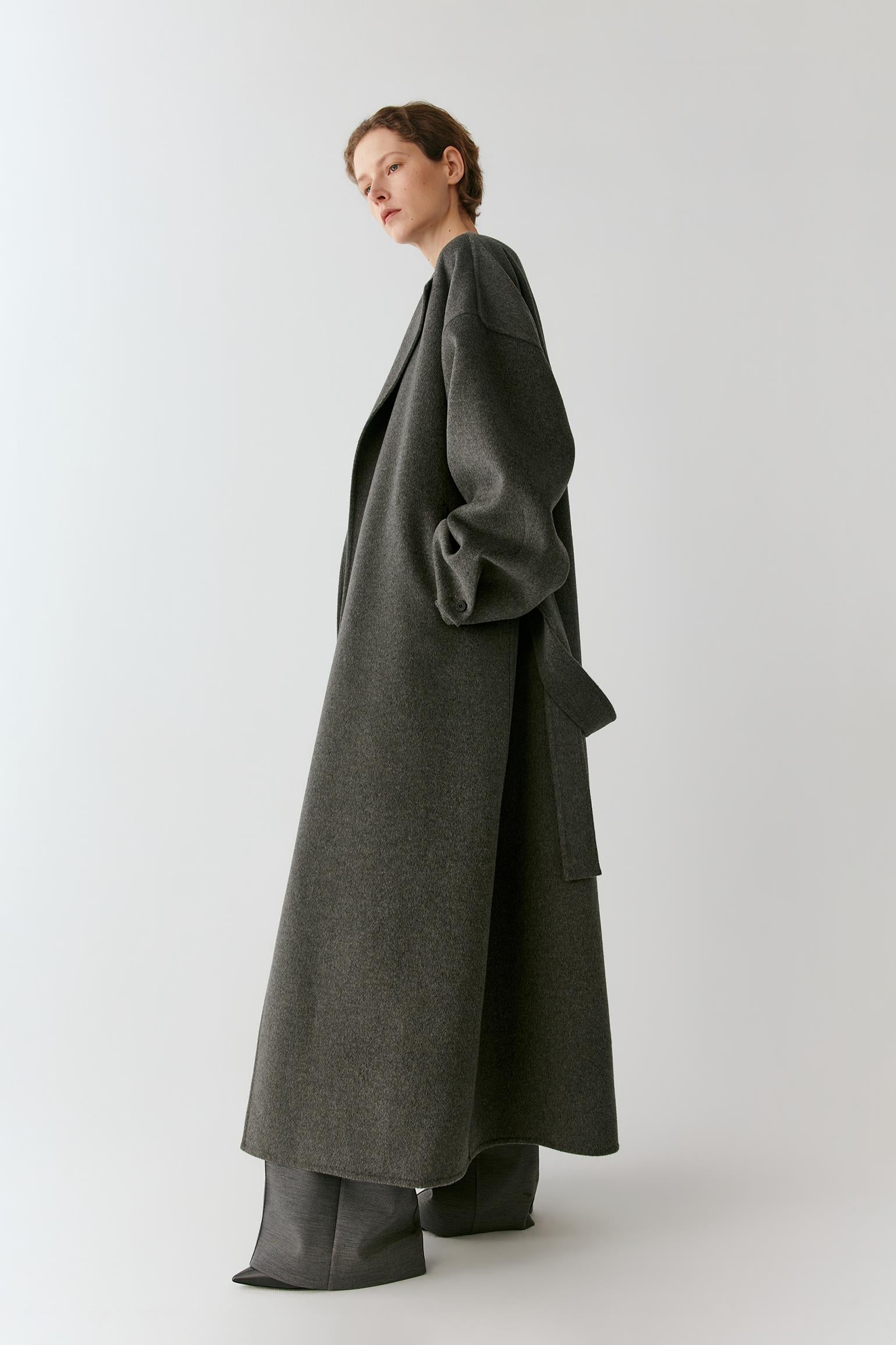 A model wearing a long, dark gray wool-blend coat with oversized draped sleeves and wide-leg trousers, photographed against a neutral light gray studio background.