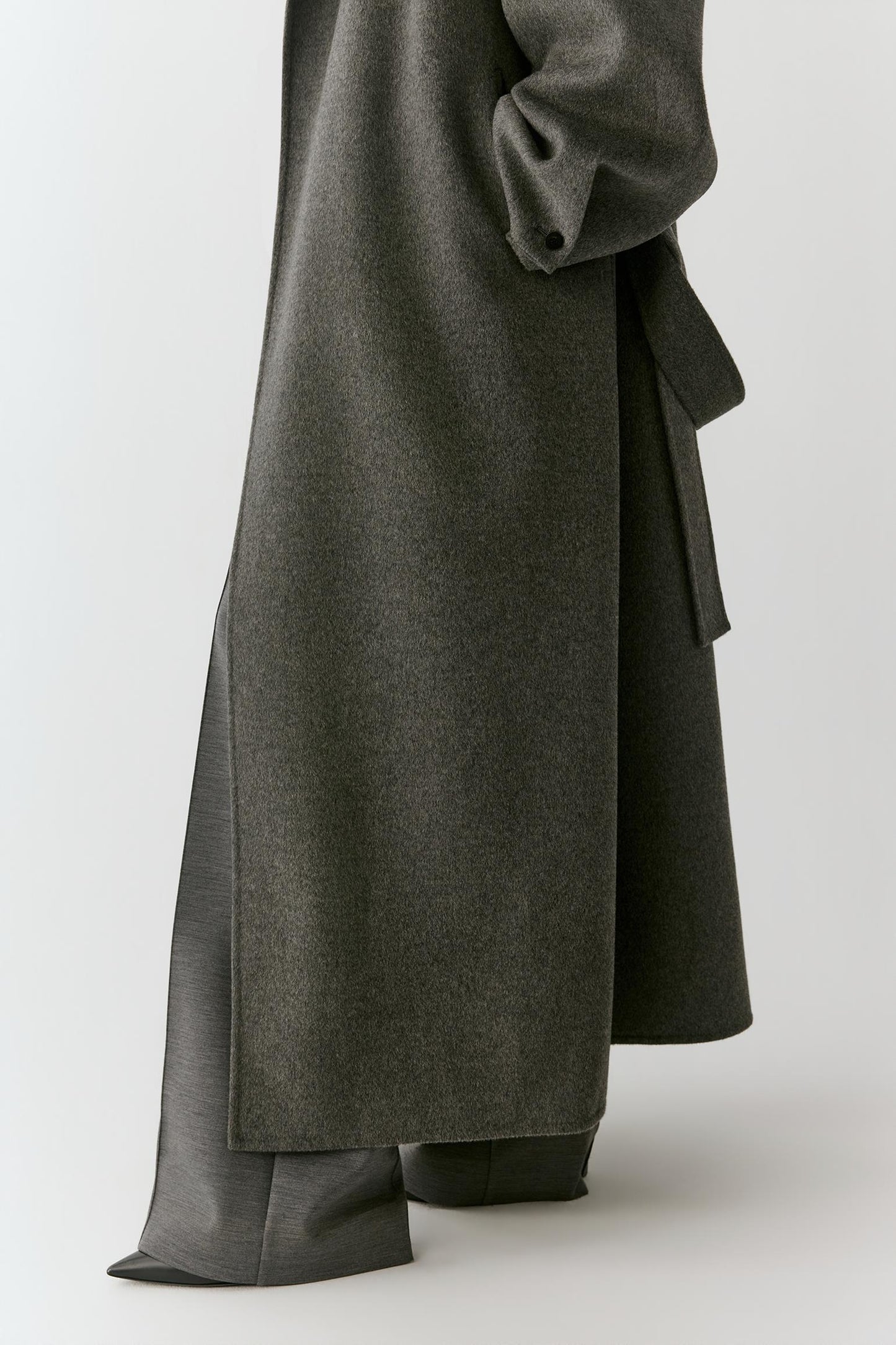 A close-up of a person wearing a long, dark gray wrap-style coat or dress with draped fabric and wide-leg pants, photographed against a light gray studio background.