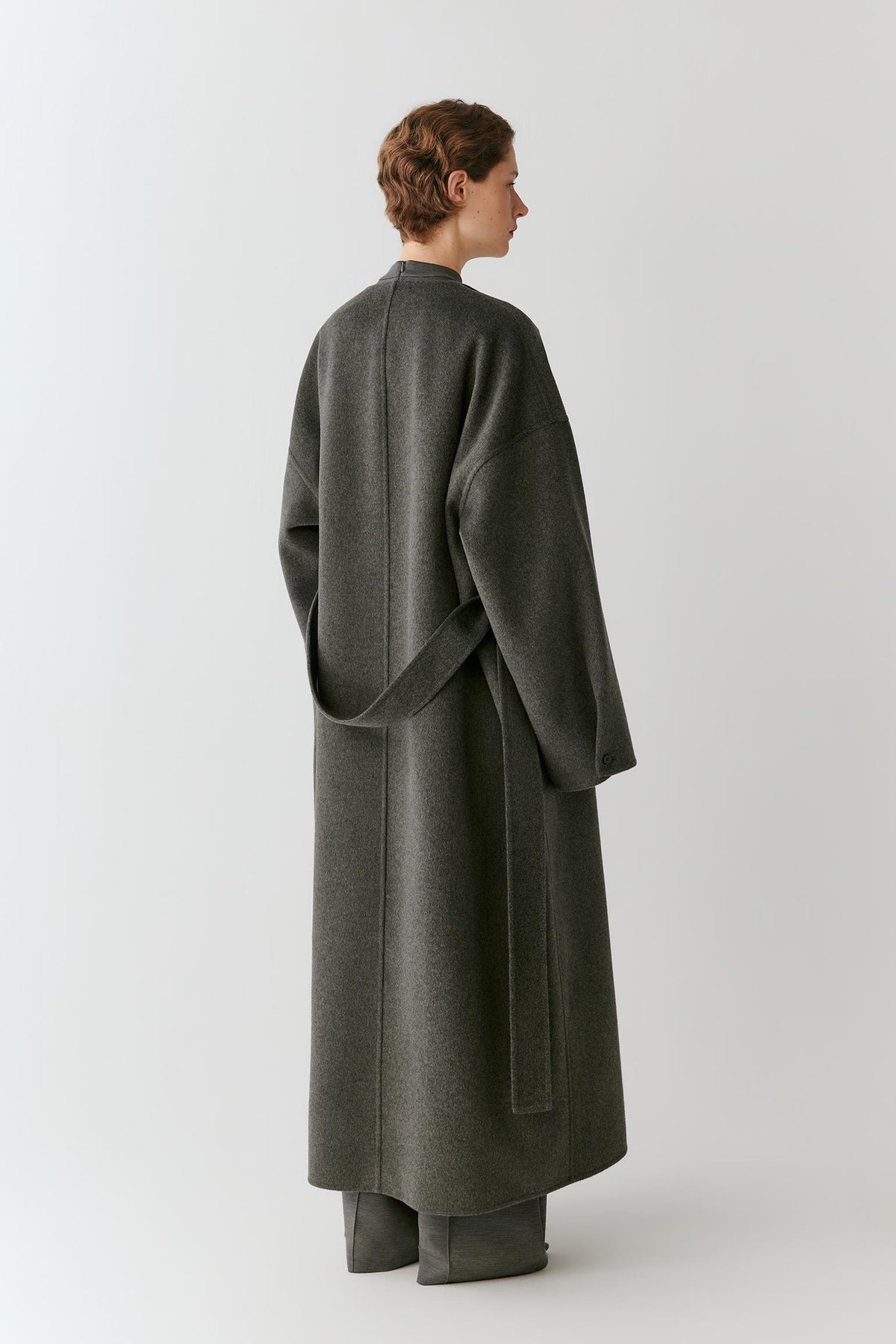 A model shown from the back wearing a long, dark gray wool coat with a matching waist tie belt, paired with matching wide-leg trousers, against a light gray studio background.