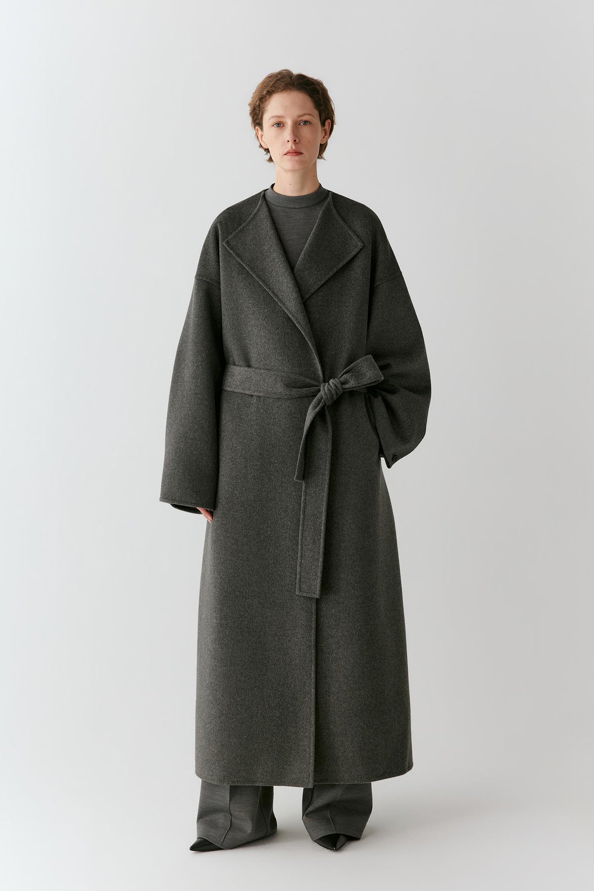 A model stands against a light gray studio background wearing a long dark charcoal gray wrap coat with a matching waist tie, paired with matching wide-leg trousers.