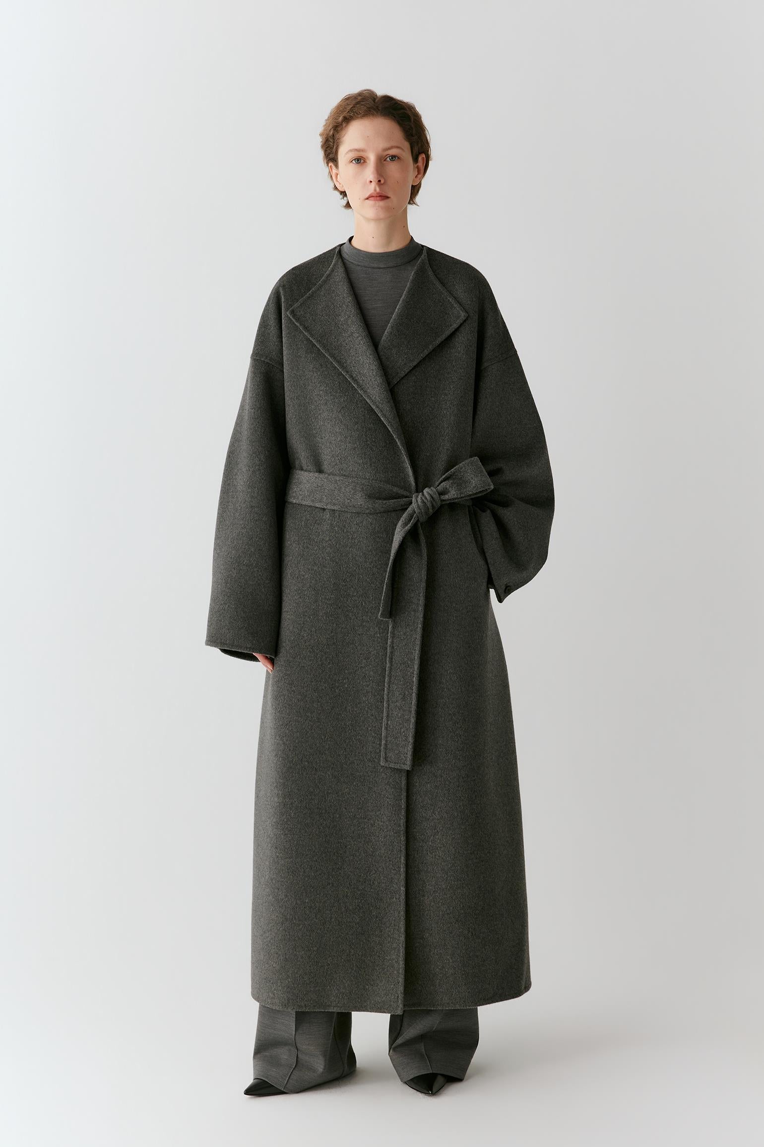 A model stands against a light gray studio background wearing a long dark charcoal gray wrap coat with a matching waist tie, paired with matching wide-leg trousers.