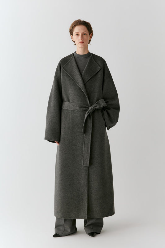 A model stands against a light gray studio background wearing a long dark charcoal gray wrap coat with a matching waist tie, paired with matching wide-leg trousers.