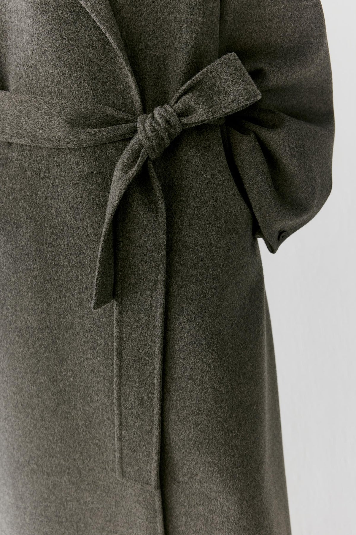 Close-up of a dark olive gray wool coat with a matching tie belt, showing the texture and fabric detail against a light gray background.