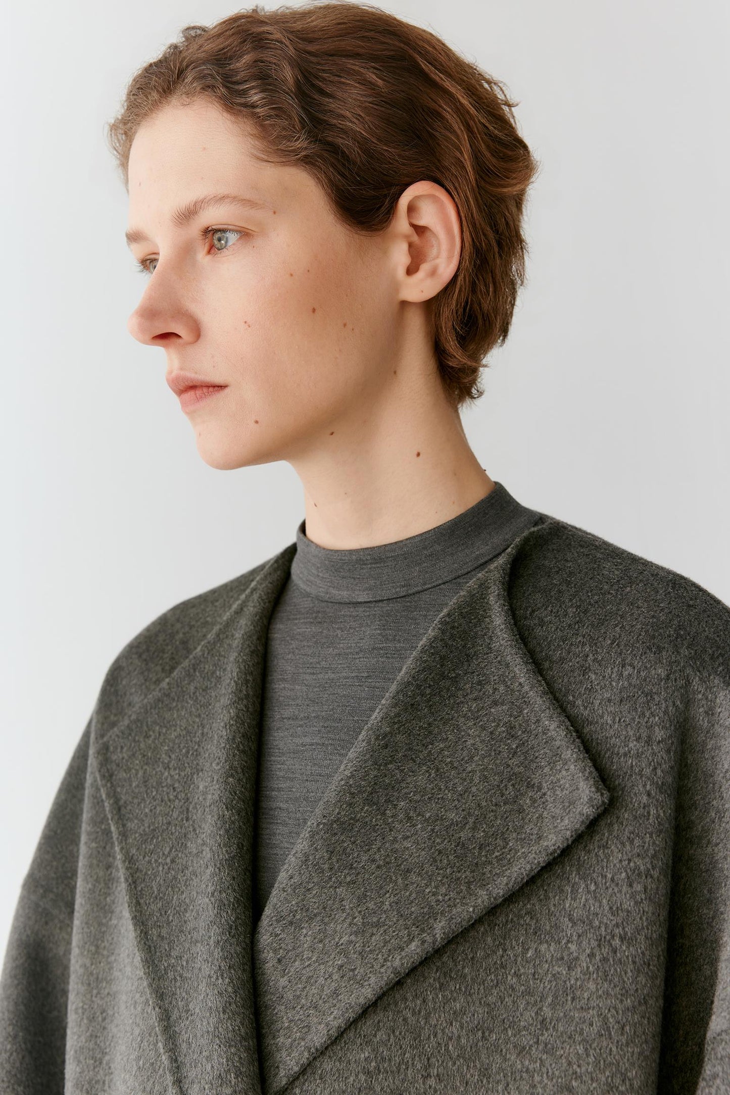 A close-up side profile of a model wearing a dark gray wool-like cape jacket over a matching gray mock-neck top, set against a plain light gray background.