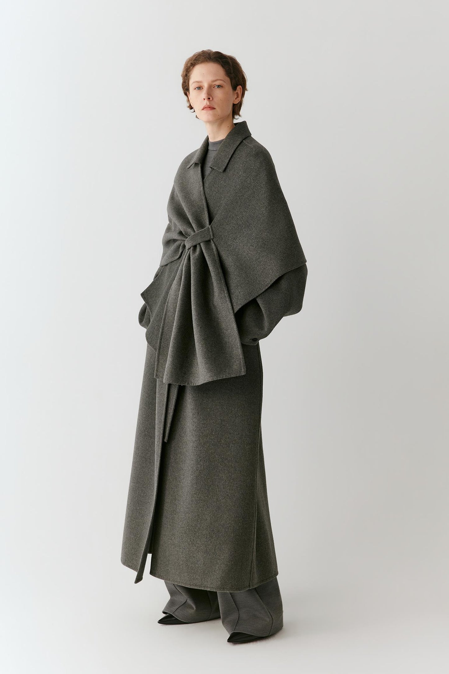 A woman wearing a dark gray wrap-style cape over a matching gray coat, photographed against a plain light gray studio background.