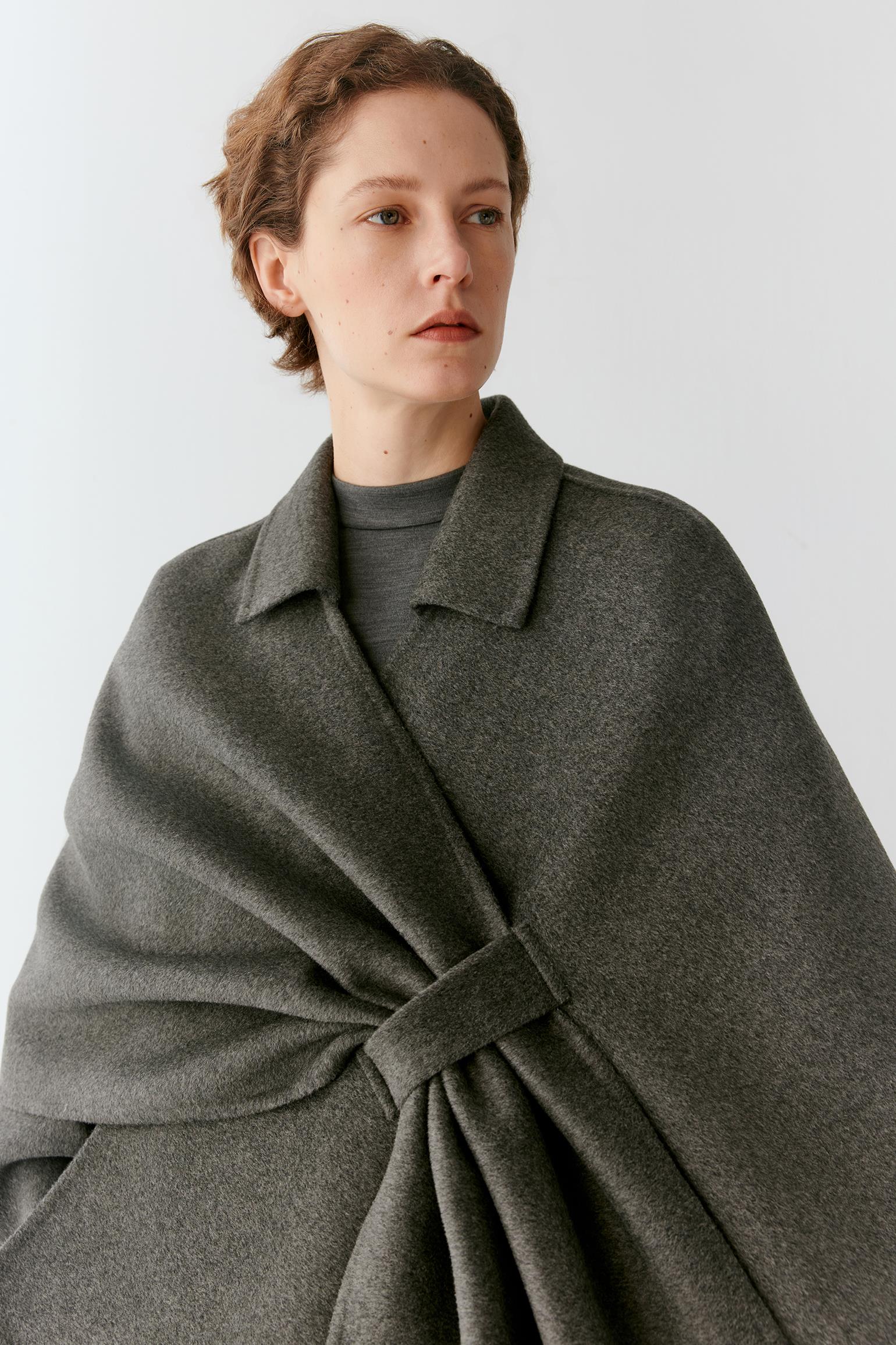 A woman wearing a dark gray wrap-style cape over a matching gray coat, photographed against a plain light gray studio background.