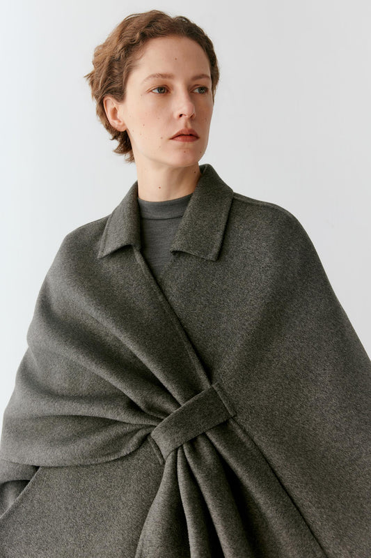 A woman wearing a dark gray wrap-style cape over a matching gray coat, photographed against a plain light gray studio background.