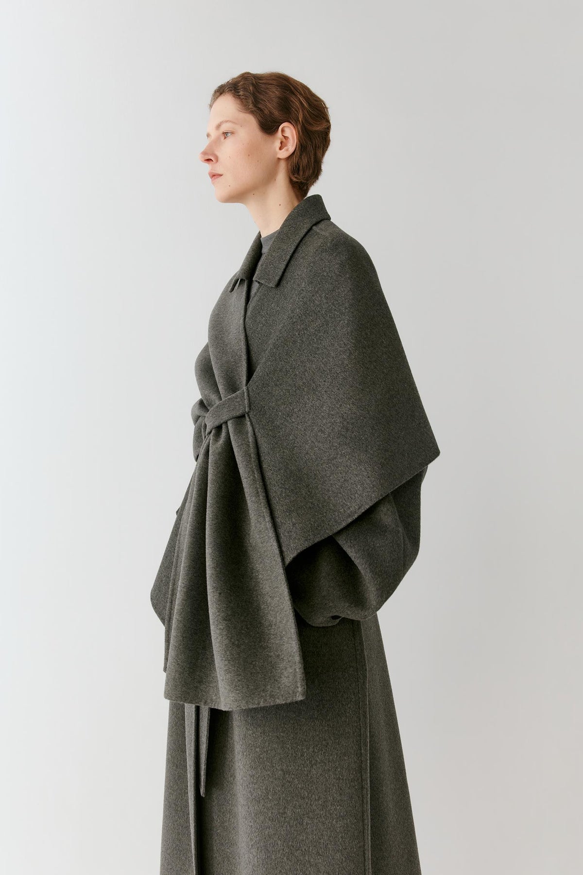 A woman wearing a dark gray wrap-style cape over a matching gray coat, photographed against a plain light gray studio background.