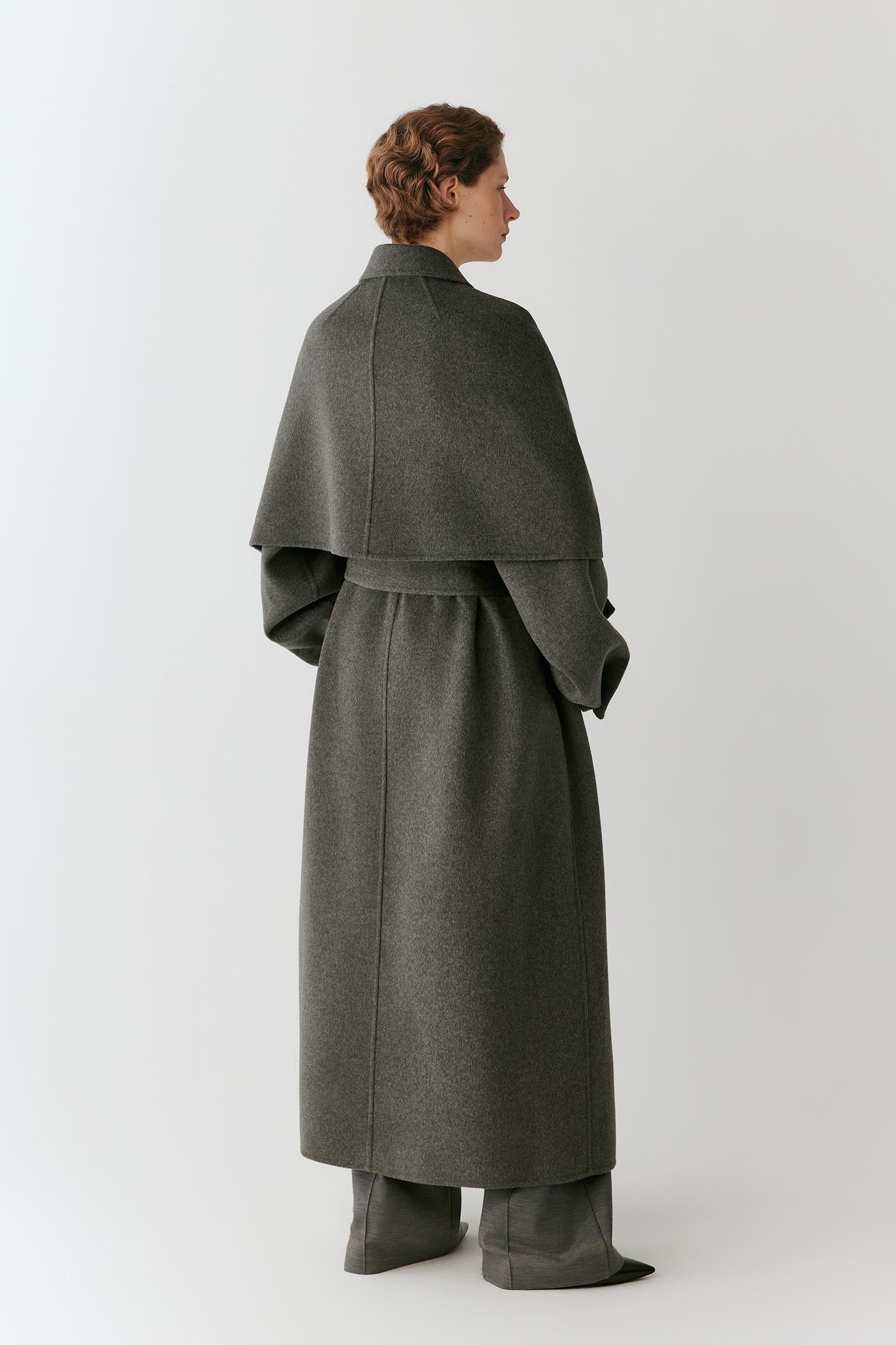 A woman wearing a dark gray wrap-style cape over a matching gray coat, photographed against a plain light gray studio background.
