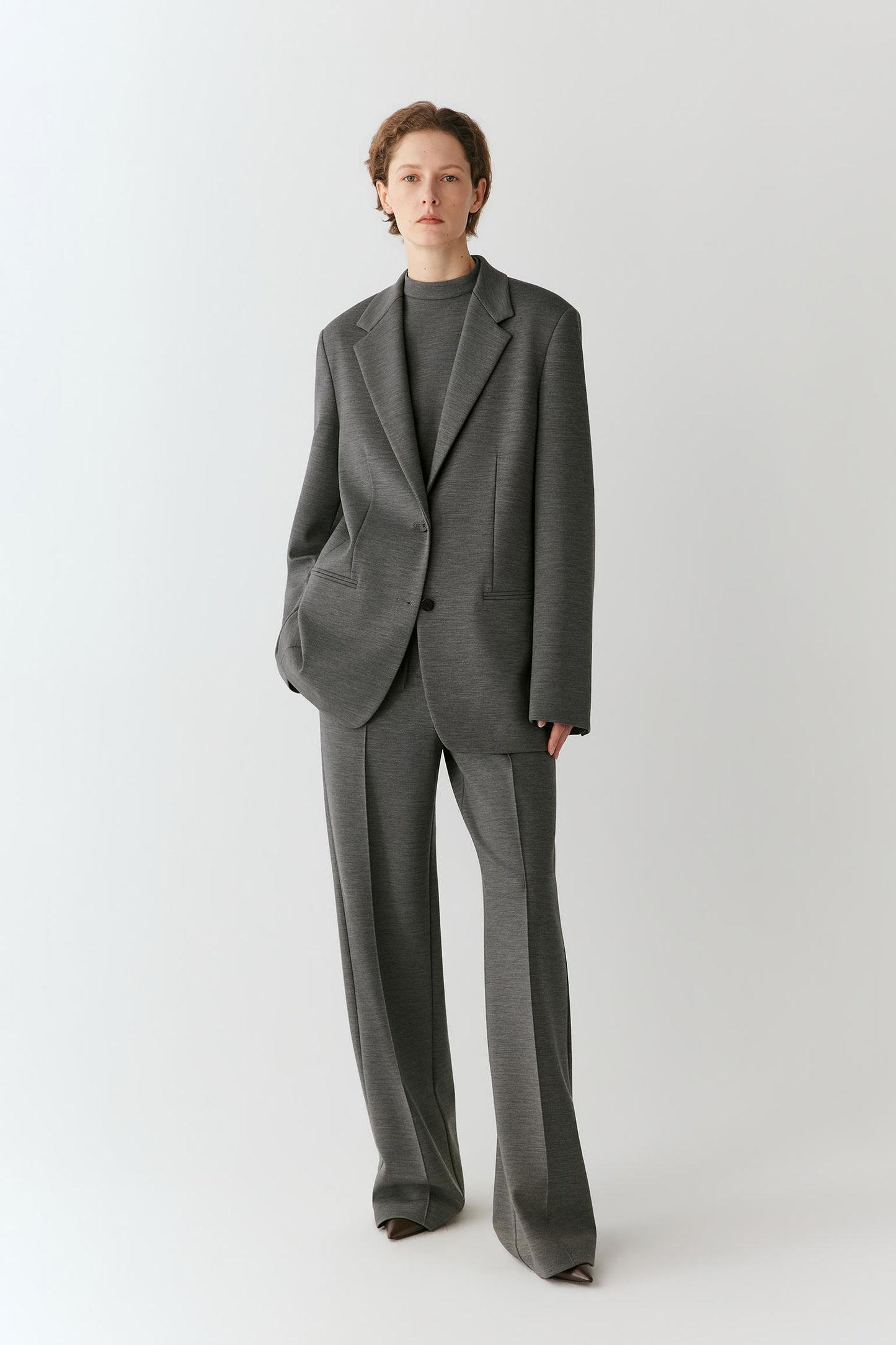 A model stands against a light gray studio background wearing a tailored gray suit with a single-breasted blazer, wide-leg trousers, and a dark gray mock-neck top.
