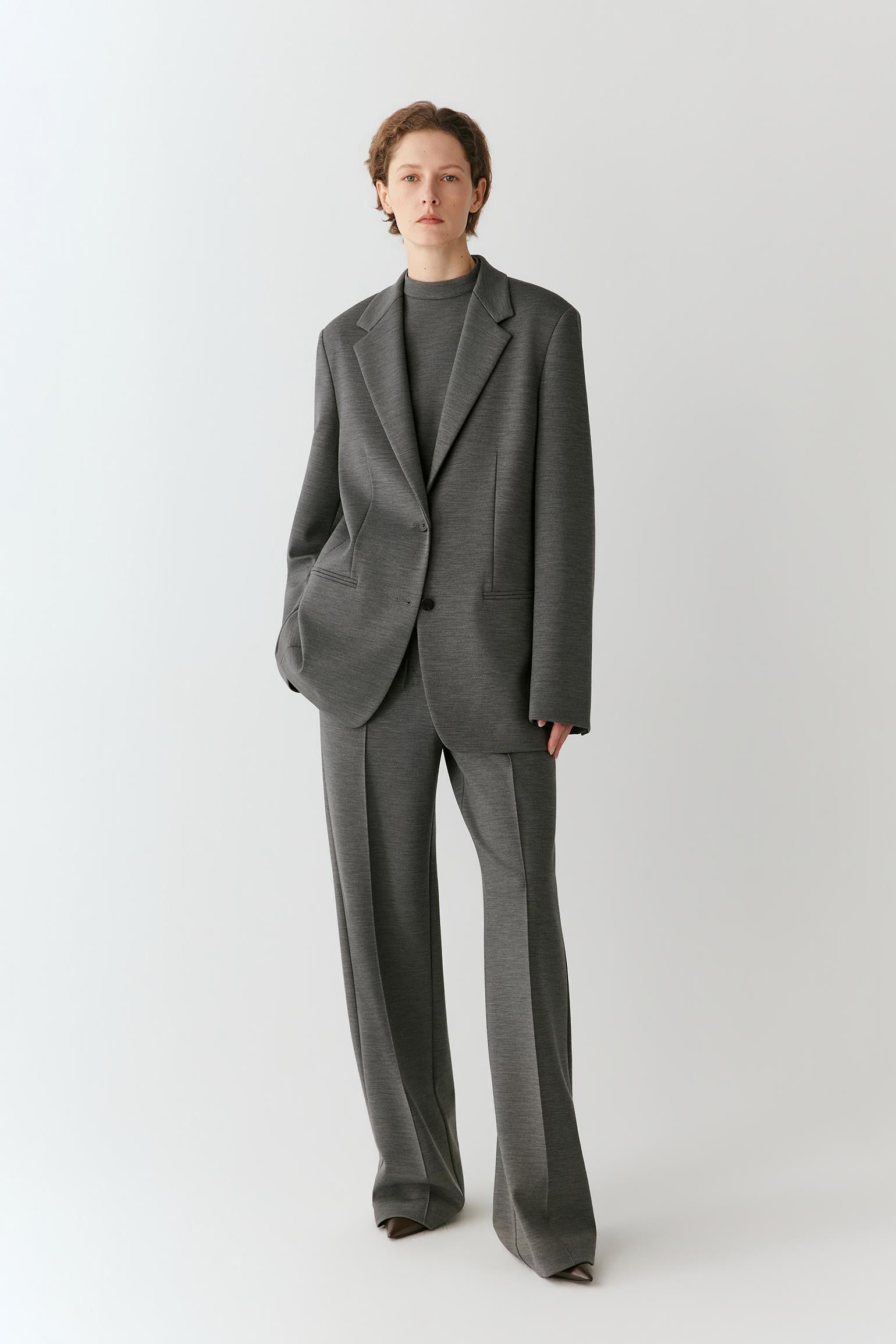 A model stands against a light gray studio background wearing a tailored gray suit with a single-breasted blazer, wide-leg trousers, and a dark gray mock-neck top.