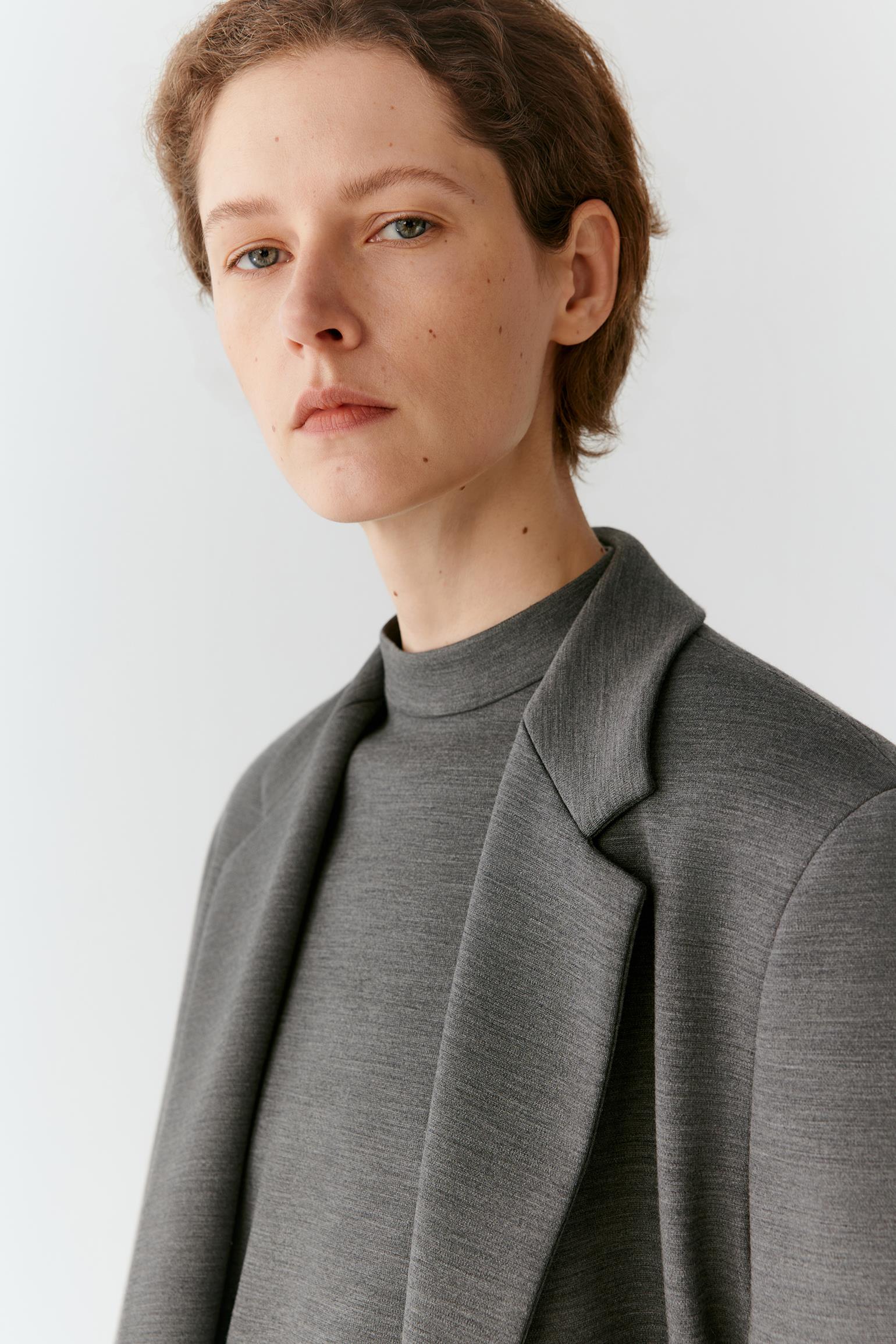 A close-up portrait of a model wearing a textured gray mock-neck top and a matching gray blazer against a light gray background.