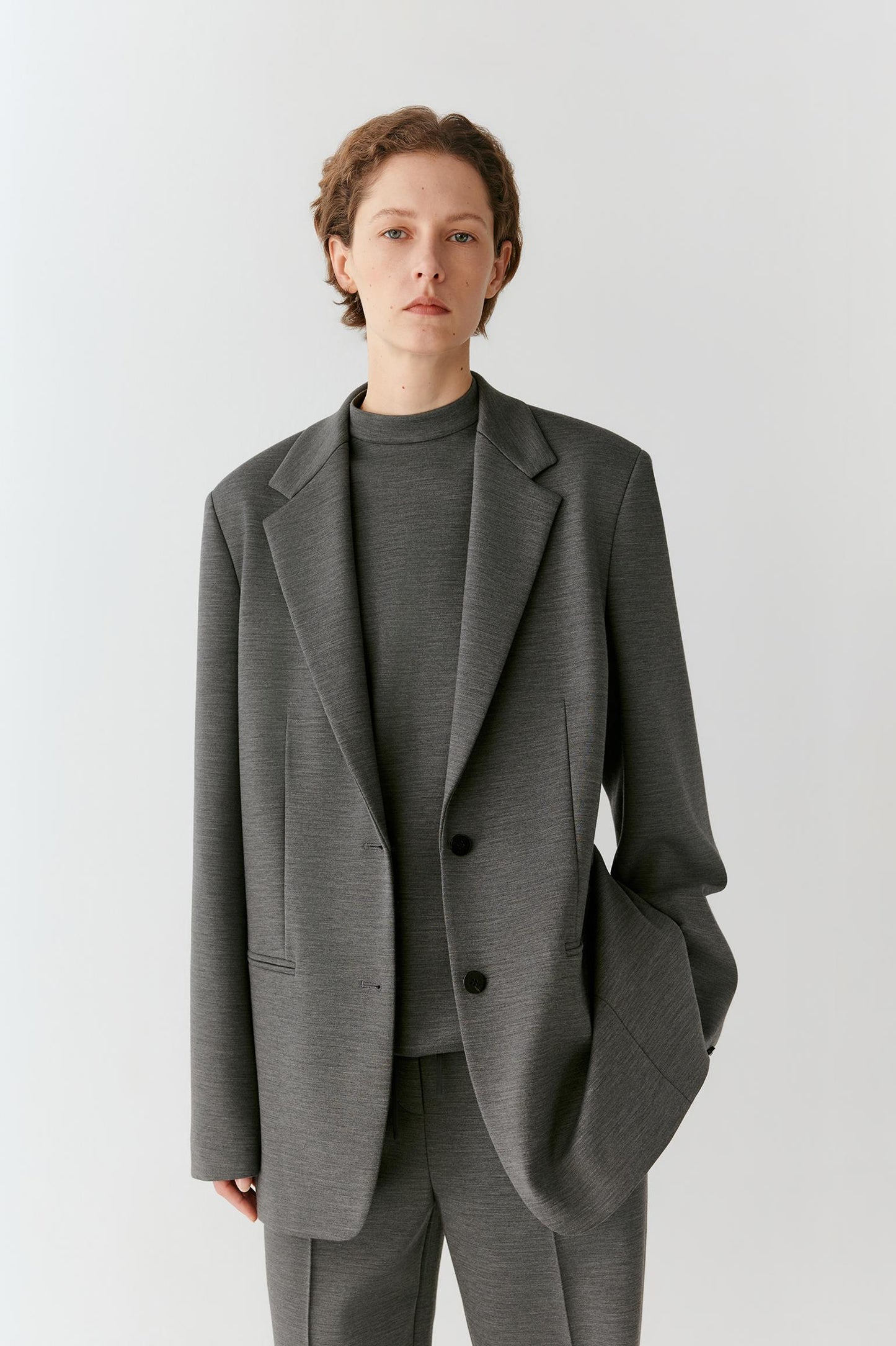 A model wearing a dark gray textured blazer over a matching gray mock-neck top and trousers, photographed against a light gray studio background.