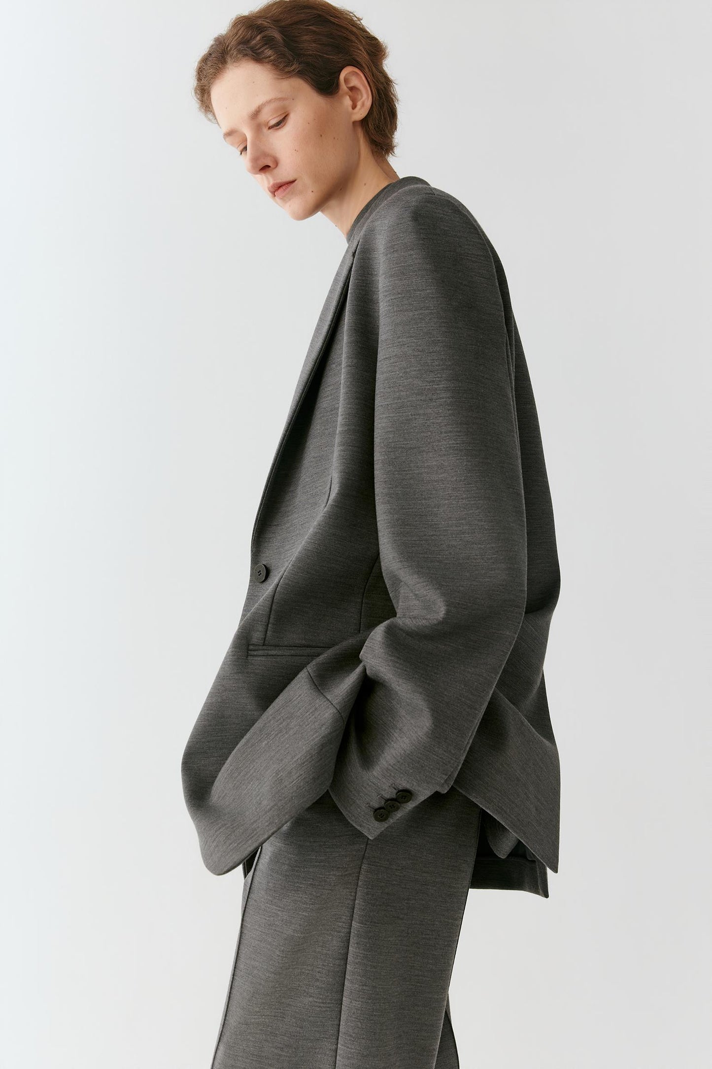 A model wearing an oversized dark gray wool-blend blazer and matching trousers, shown from a side profile against a light gray studio background.