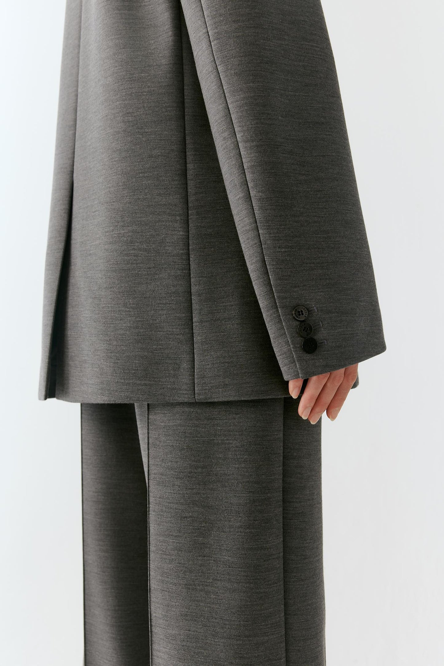 Close-up of a person wearing a dark gray tailored suit with wide-leg trousers and a matching blazer, showing the sleeve with three buttons and part of the hand.