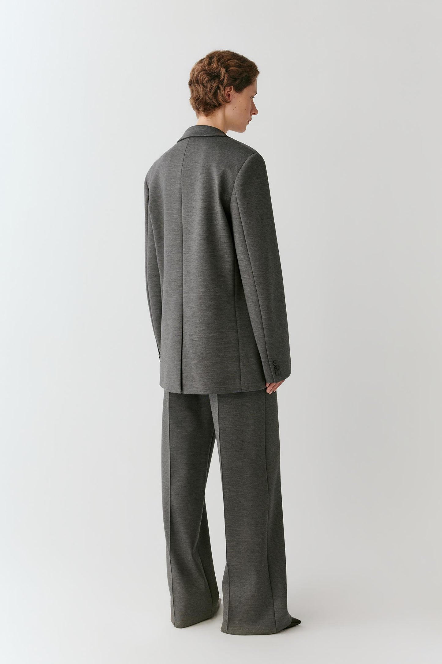 A model shown from the back wearing a dark gray two-piece suit with a blazer and wide-leg trousers, photographed against a neutral light gray studio background.