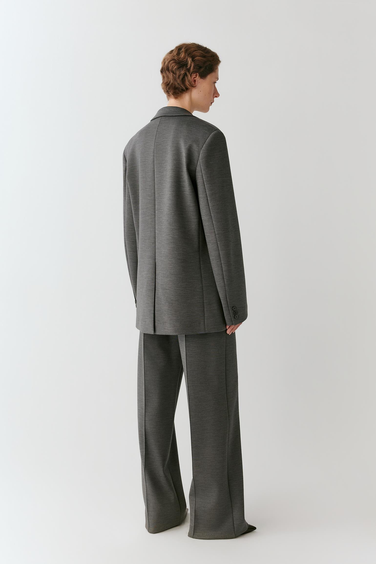 A model shown from the back wearing a dark gray two-piece suit with a blazer and wide-leg trousers, photographed against a neutral light gray studio background.
