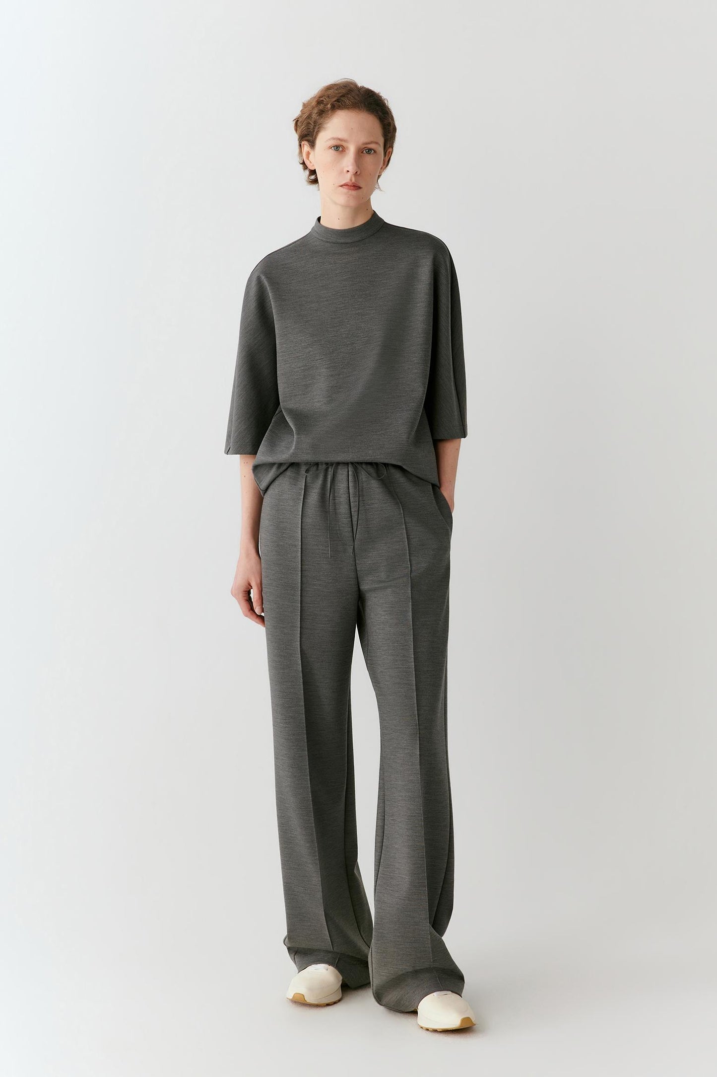 A model stands against a light gray studio background wearing a dark gray mock-neck top and matching wide-leg trousers, paired with off-white slip-on shoes.