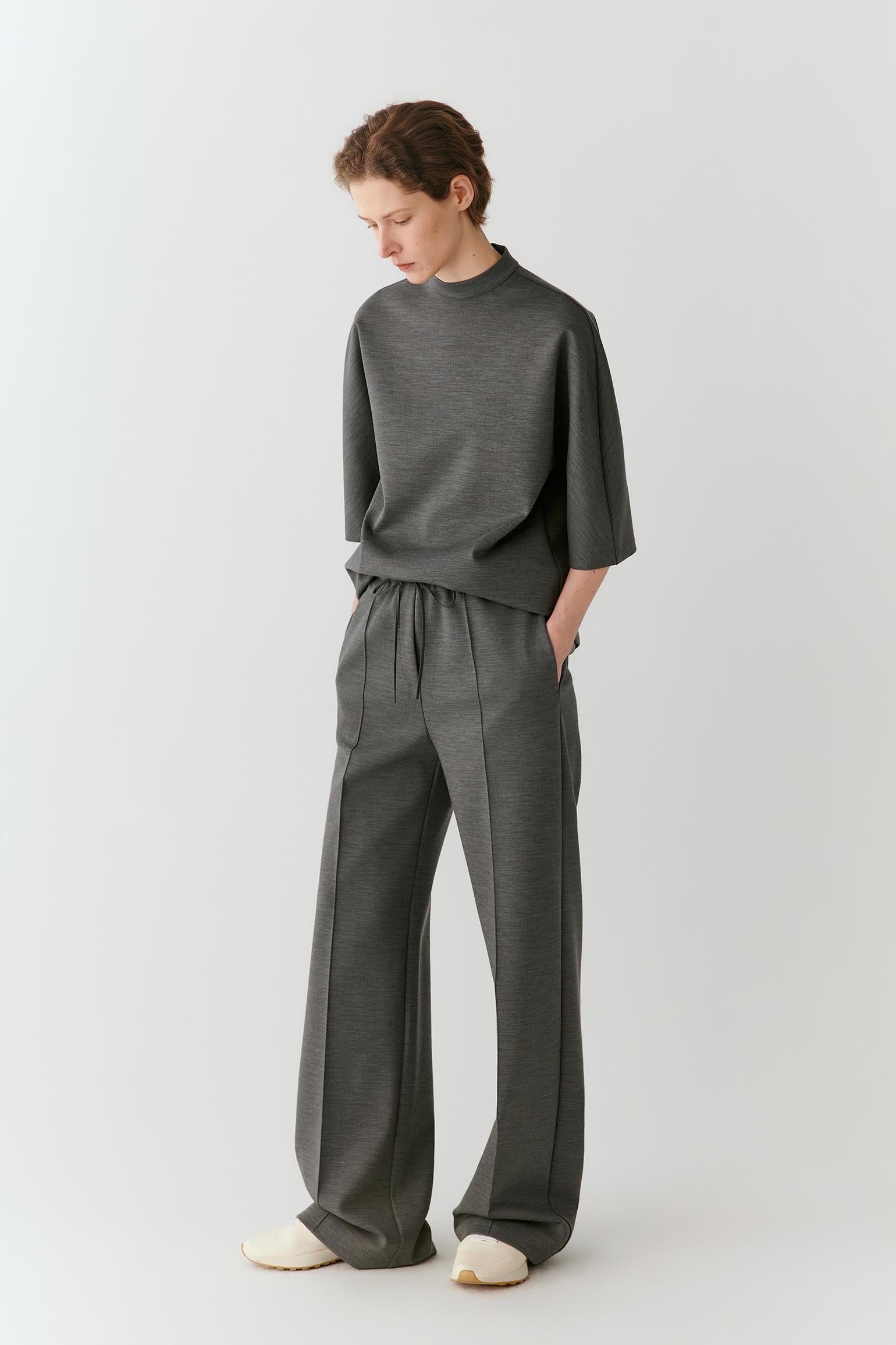 A model wearing a dark gray mock-neck knit top and matching wide-leg drawstring trousers, standing against a light gray studio background.