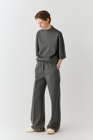 A model wearing a dark gray mock-neck knit top and matching wide-leg drawstring trousers, standing against a light gray studio background.