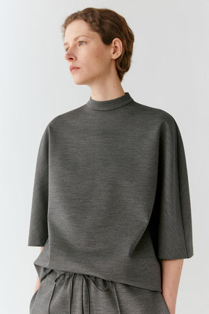 A model wearing a dark gray heathered mock neck top with elbow-length sleeves and matching gray drawstring trousers, photographed against a light gray studio background.