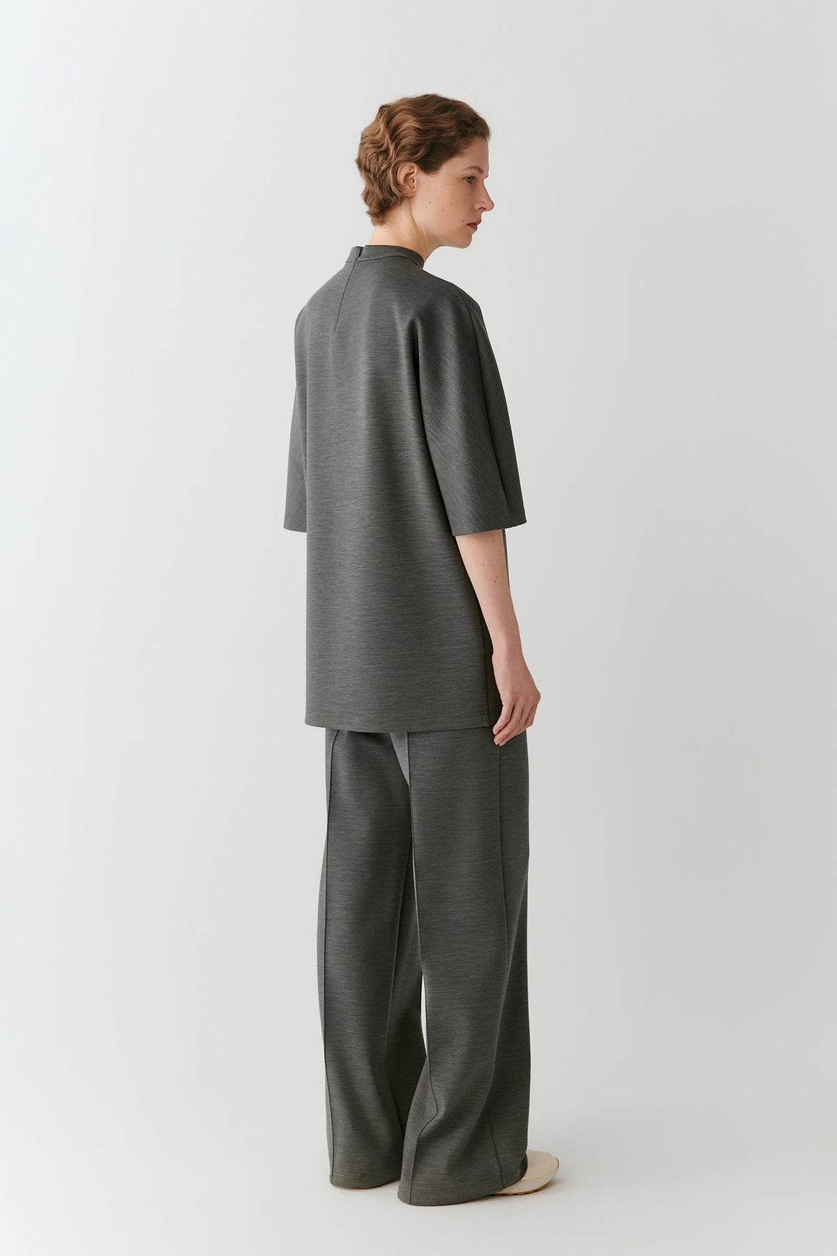 A model shown from the back wearing a dark gray textured two-piece outfit consisting of a short-sleeve top and wide-leg trousers, photographed against a clean light gray studio background.