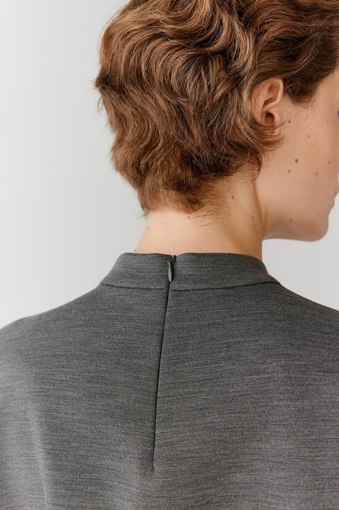 Close-up back view of a person with short wavy brown hair wearing a dark gray ribbed knit top or sweater, showing the center back zipper and collar.