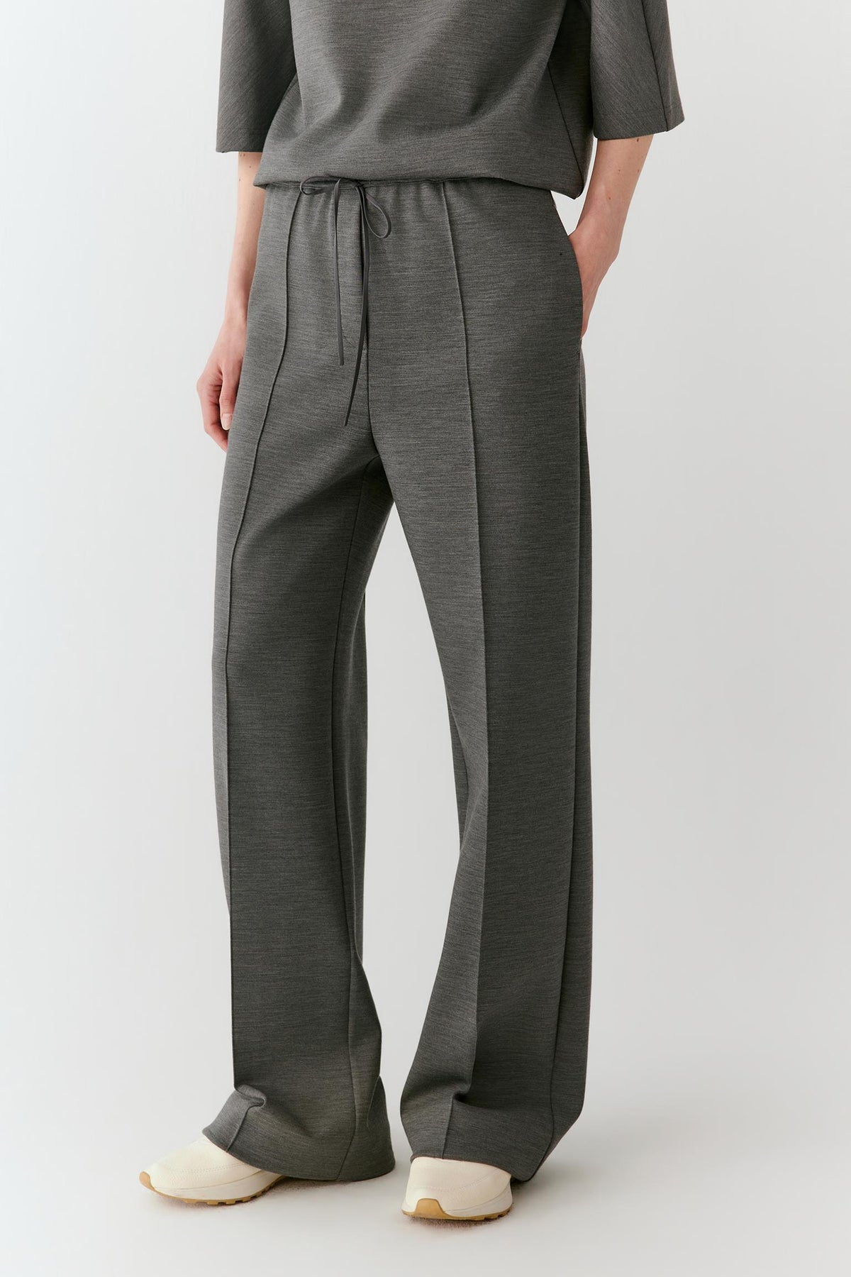 A person wearing dark gray wide-leg drawstring pants with a matching gray top and cream-colored shoes, shown from the waist down against a light gray studio background.