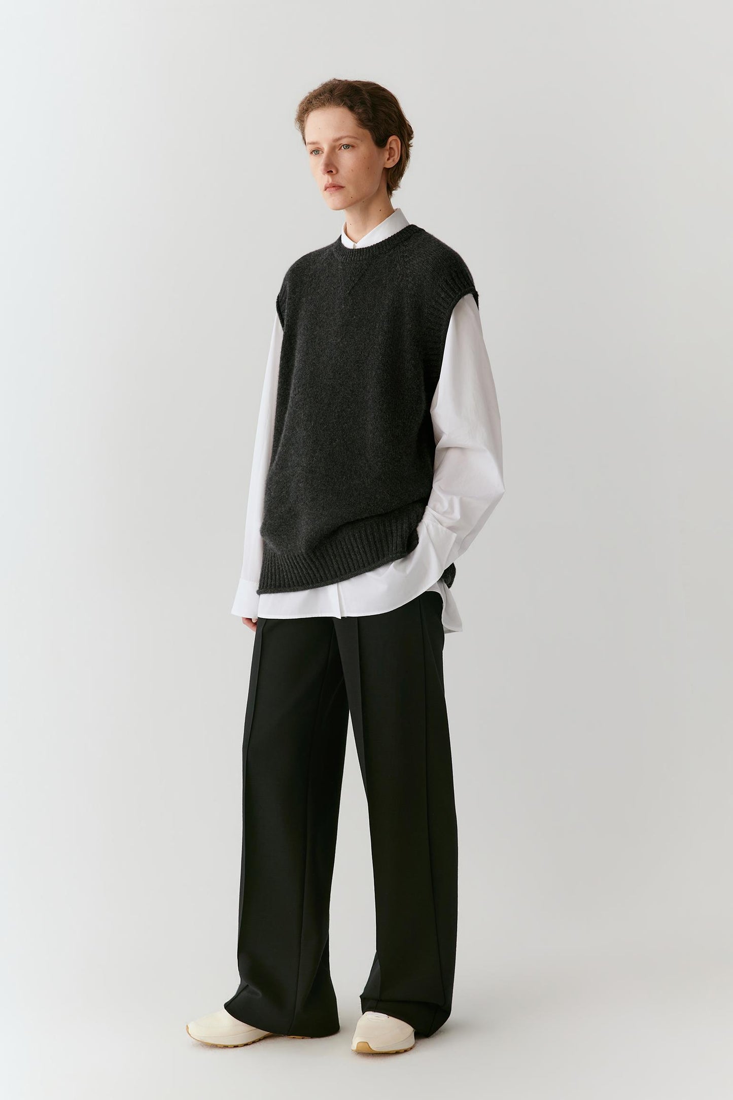 A model stands against a light gray studio background wearing a dark charcoal sleeveless knit sweater vest over a white long-sleeve shirt, paired with wide-leg black trousers and off-white slip-on shoes.