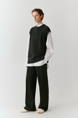 A model stands against a light gray studio background wearing a dark charcoal sleeveless knit sweater vest over a white long-sleeve shirt, paired with wide-leg black trousers and off-white slip-on shoes.