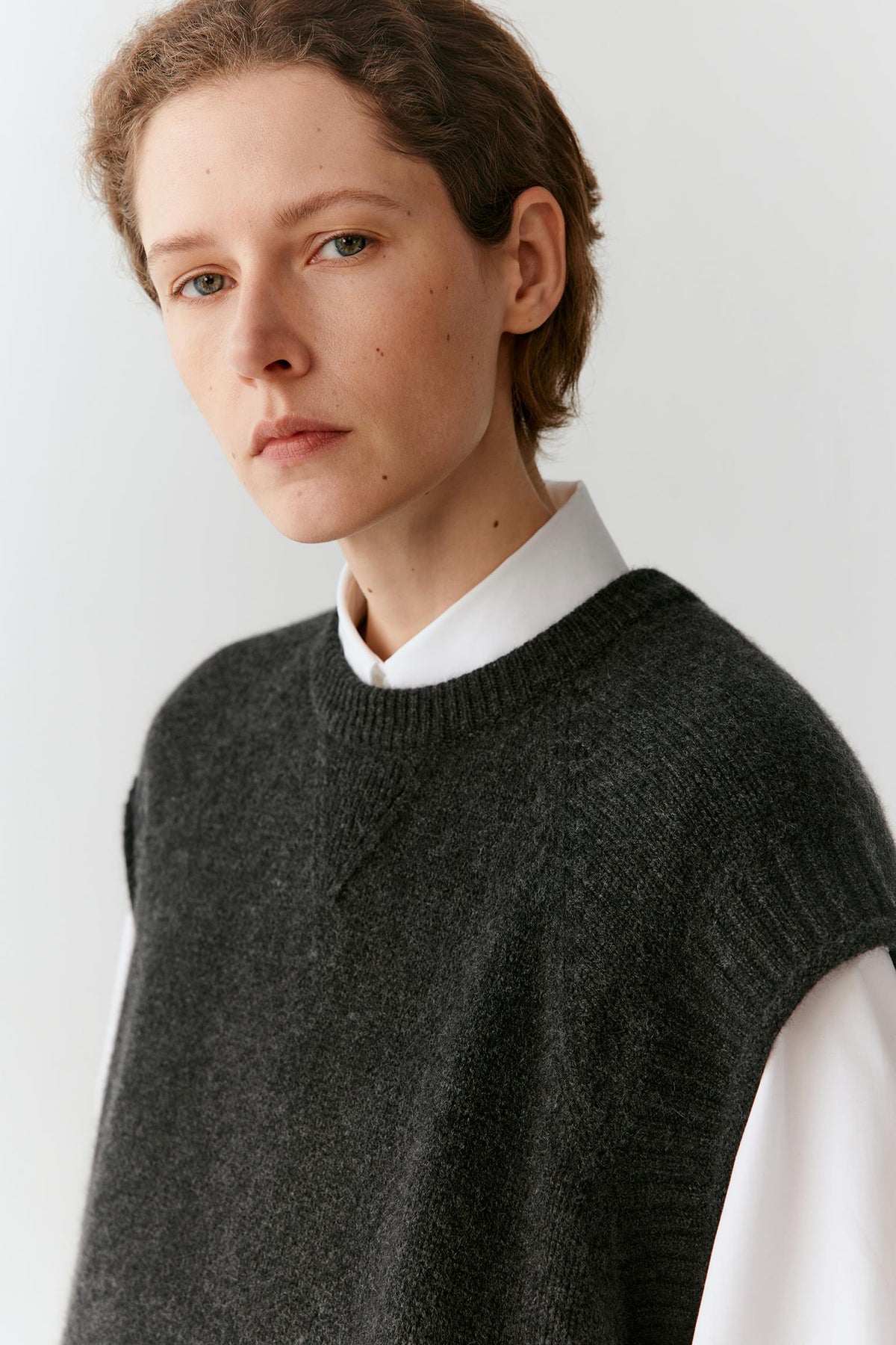 A close-up portrait of a model wearing a dark charcoal gray ribbed knit sweater vest over a white collared shirt, set against a plain light gray background.