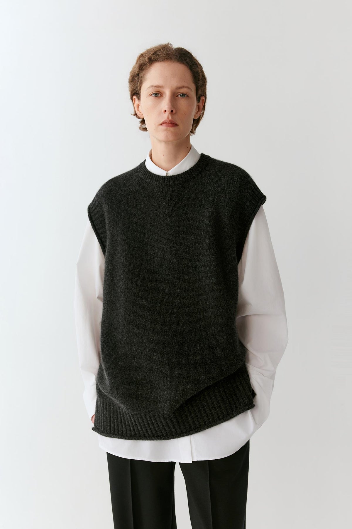 A model wearing a dark charcoal gray sleeveless knit sweater vest over a white collared shirt and black trousers, photographed against a clean white studio background.