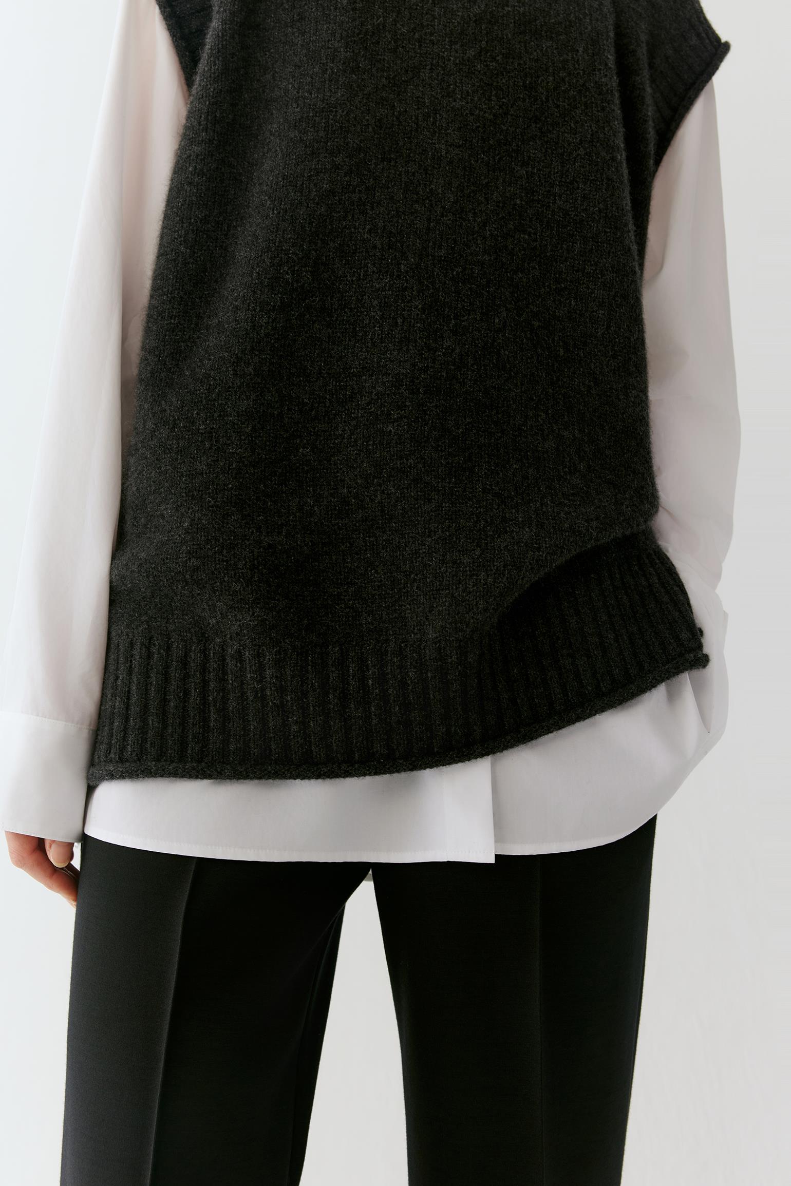A close-up of a person wearing a dark charcoal knit sleeveless sweater vest over a white long-sleeve shirt and black pants.