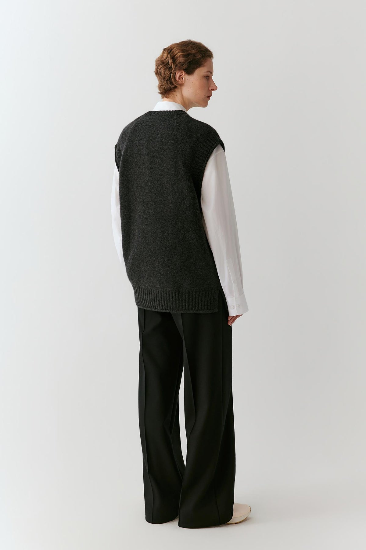 A model shown from the back wearing a dark charcoal gray ribbed knit sweater vest over a white collared shirt and black wide-leg trousers, against a plain light gray studio background.