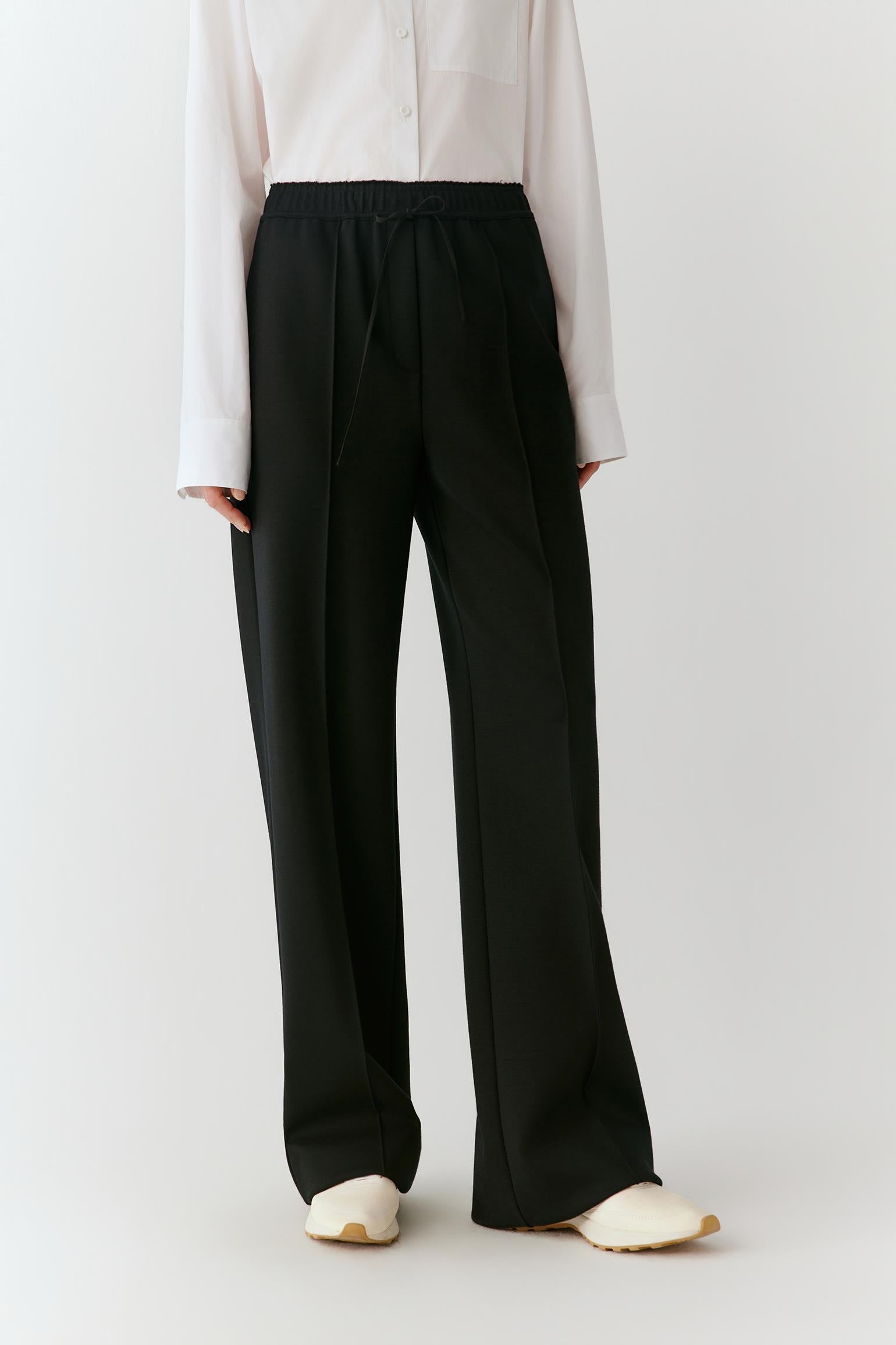 A model wearing black wide-leg trousers with an elastic waistband, paired with a white long-sleeve blouse and cream-colored slip-on shoes, photographed against a light gray studio background.