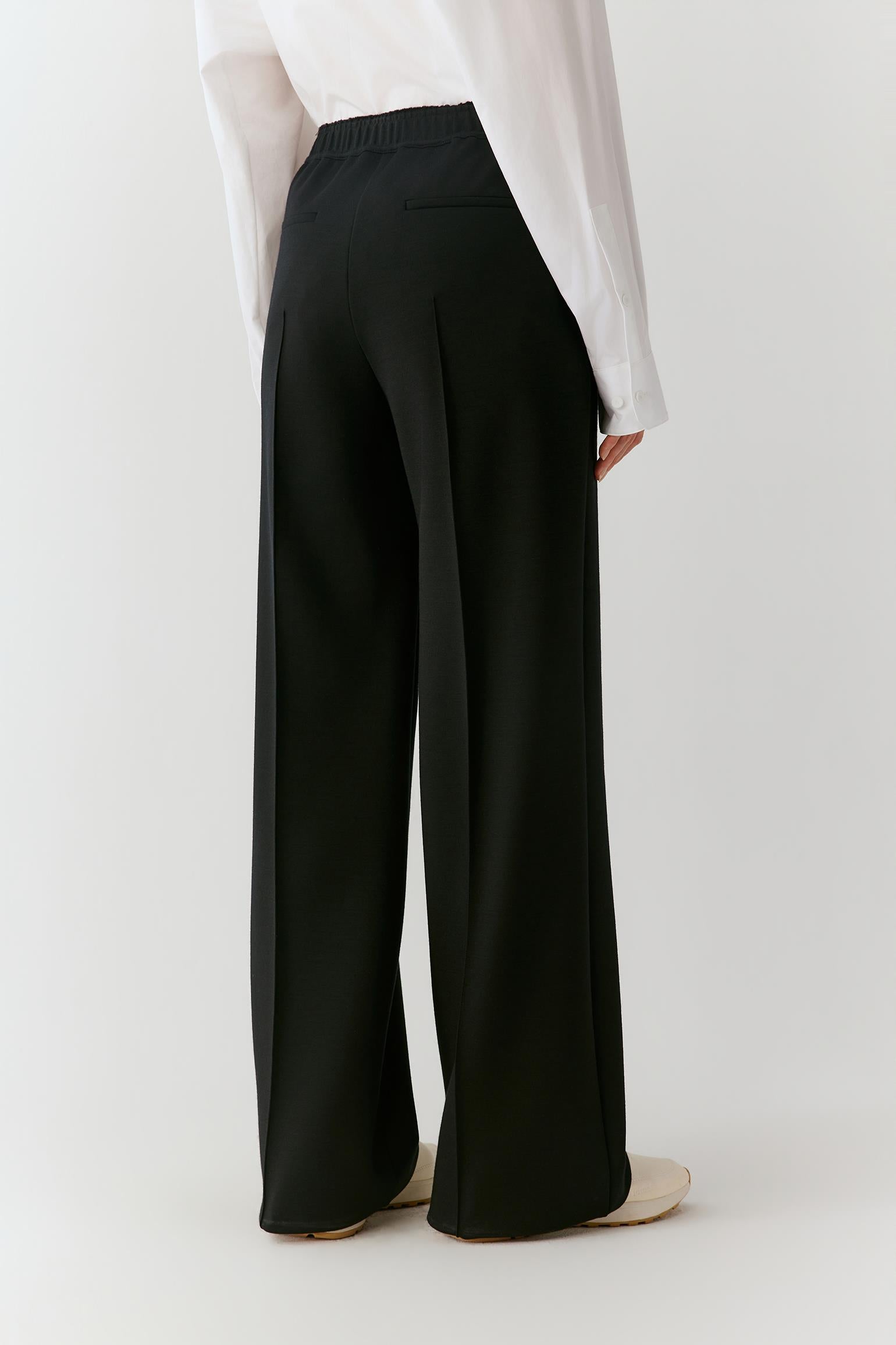 Black wide-leg trousers shown from the back on a model, paired with a white long-sleeve blouse and beige flat shoes against a light gray studio background.