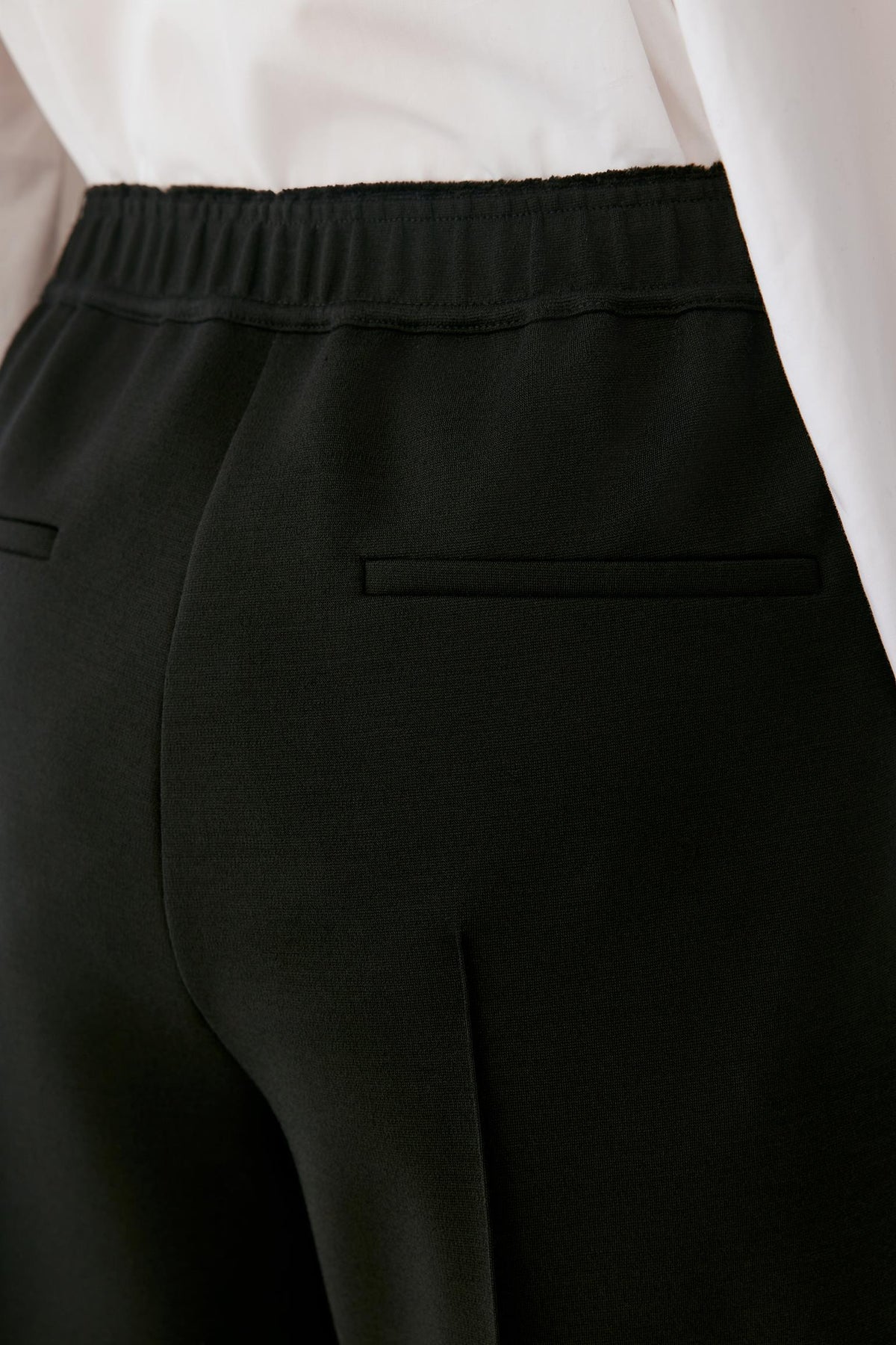 Close-up of a person wearing black trousers with an elastic waistband and a welt pocket, paired with a white top.